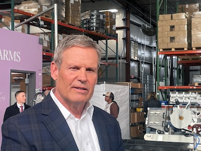 A close up of a man wearing a dark suit and a white shirt with a warehouse and two people standing in the background.