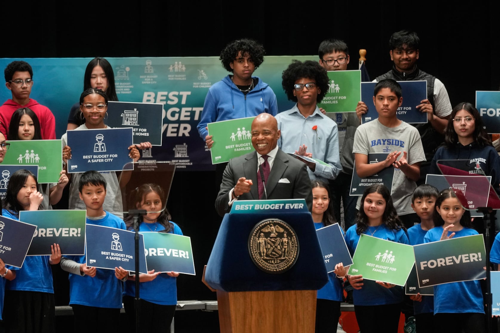 A man with a gray suit jacket stands at a podium surrounded with students holding signs.