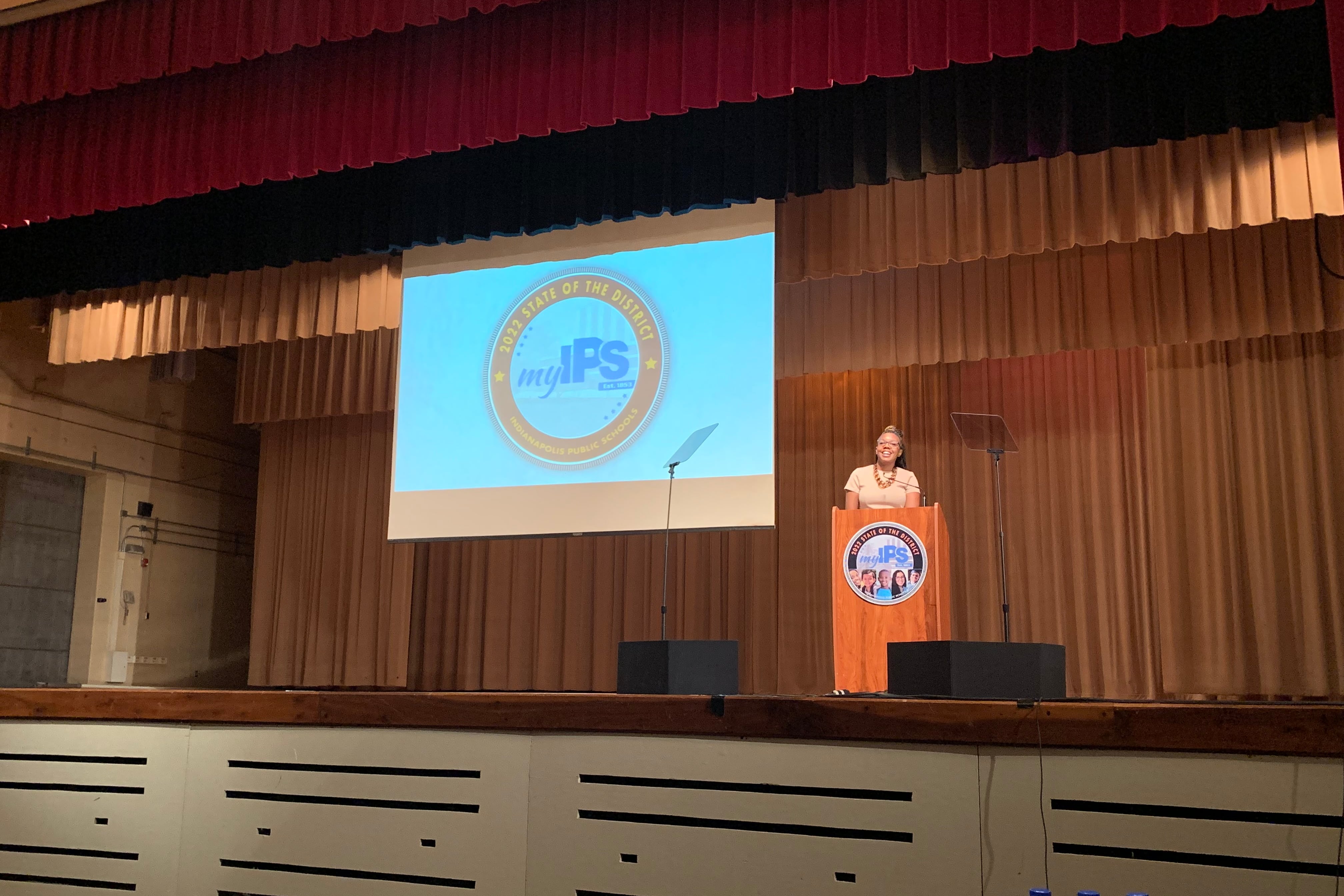 A woman stands in front of a podium on a stage. In the background is a screen with a logo that reads, “My IPS.”