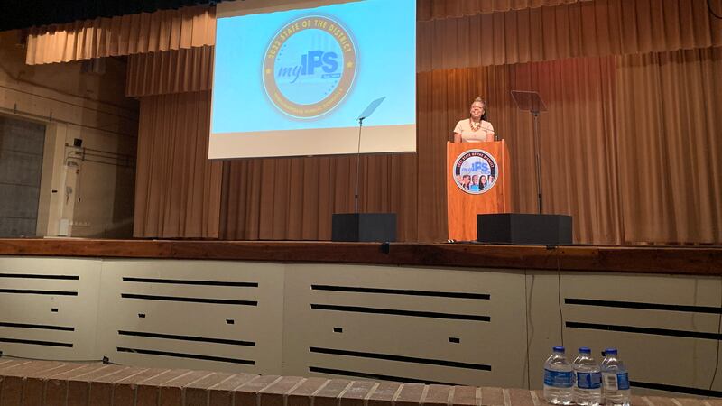 A woman stands in front of a podium on a stage. In the background is a screen with a logo that reads, “My IPS.”