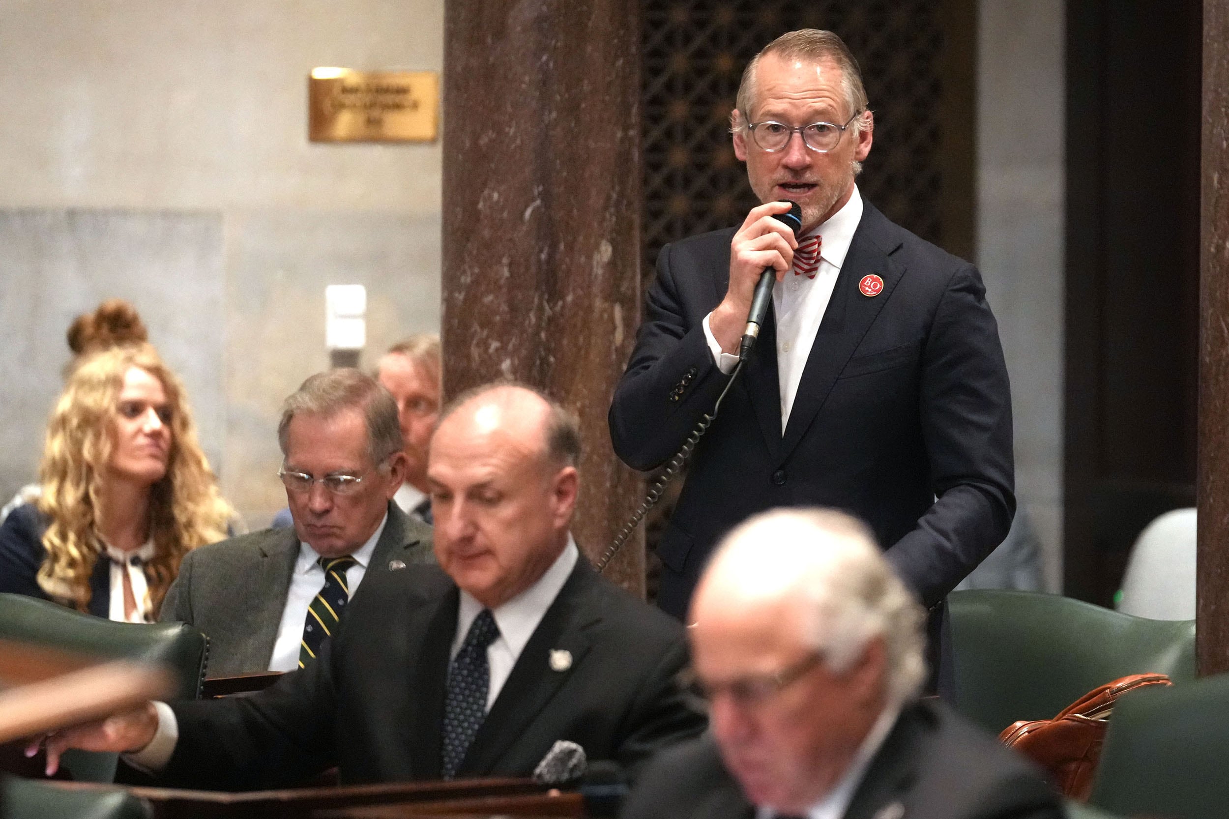 A photograph of a white man in a suit standing up in a large statehouse floor conference room speaking into a microphone with five other people in business clothes sitting around him.