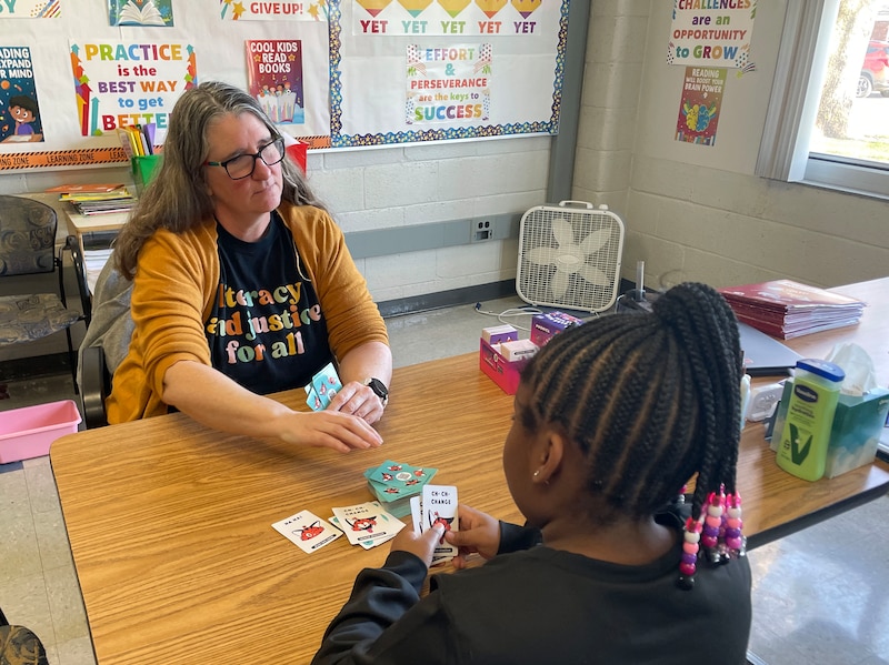 A white educator in a yellow sweater sits across the table from a young Black student with long, braided hair in a classroom.