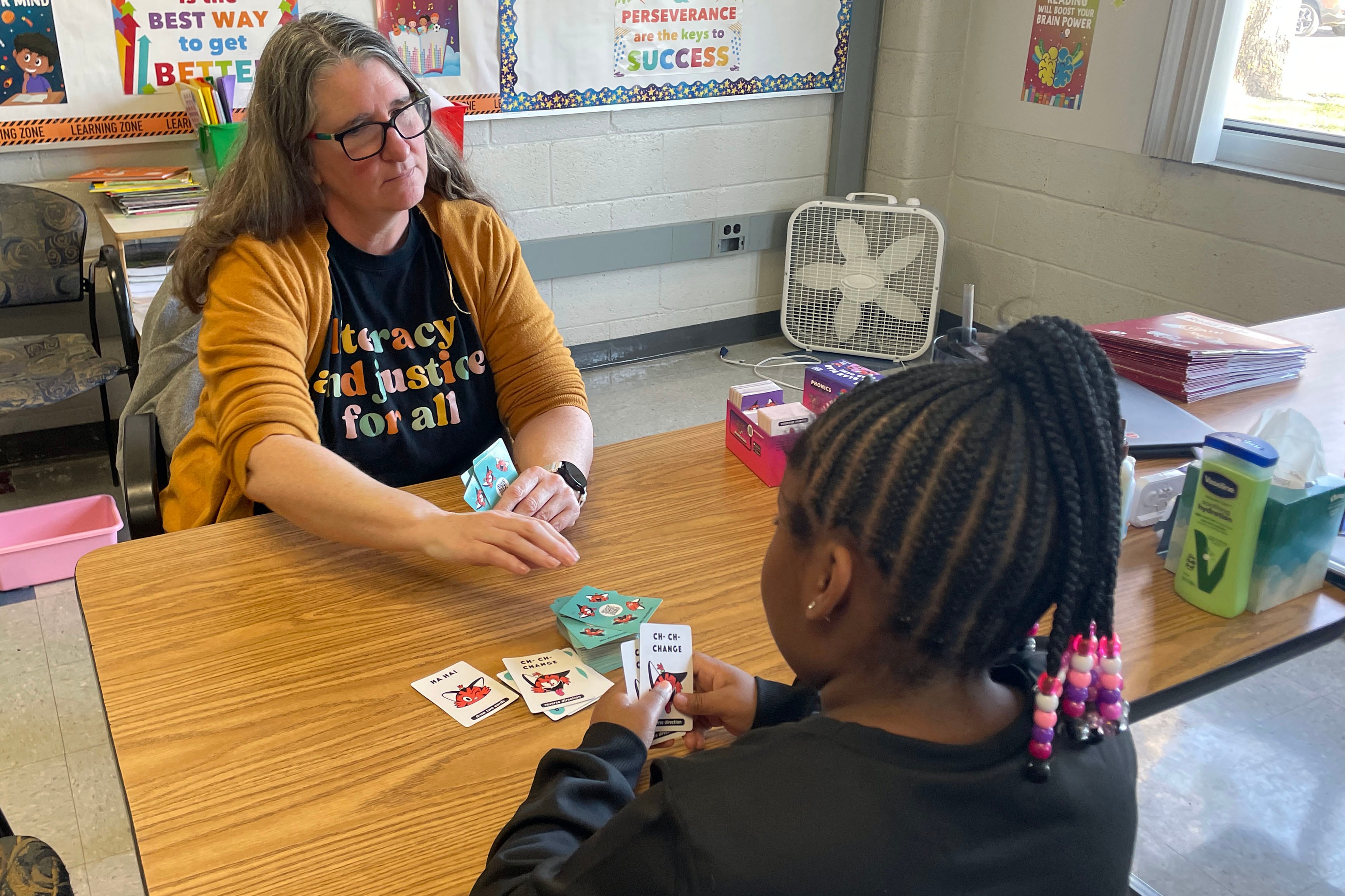 A white educator in a yellow sweater sits across the table from a young Black student with long, braided hair in a classroom.