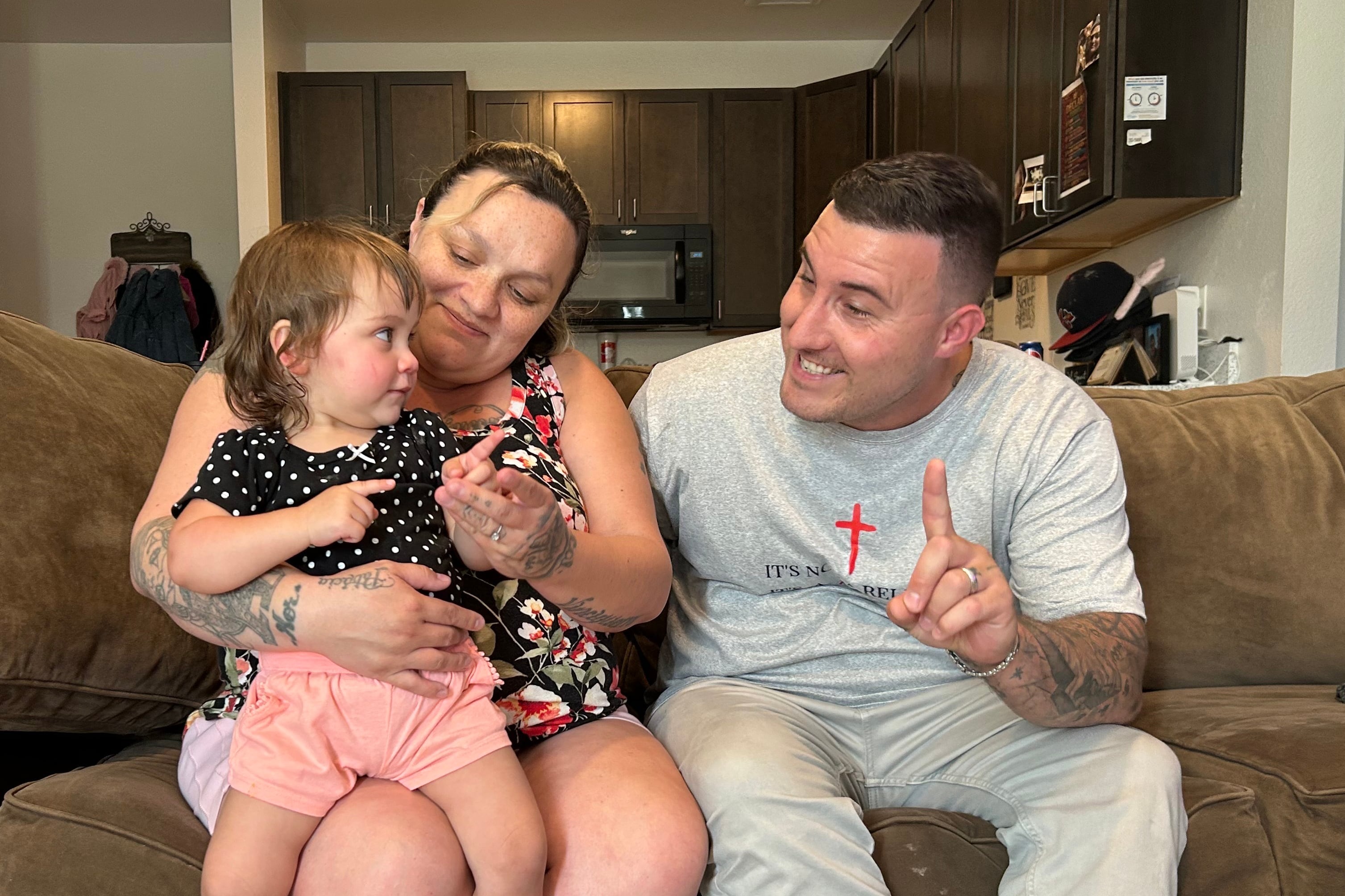 A mom and dad look at their one-year-old daughter while sitting on the couch in their home.