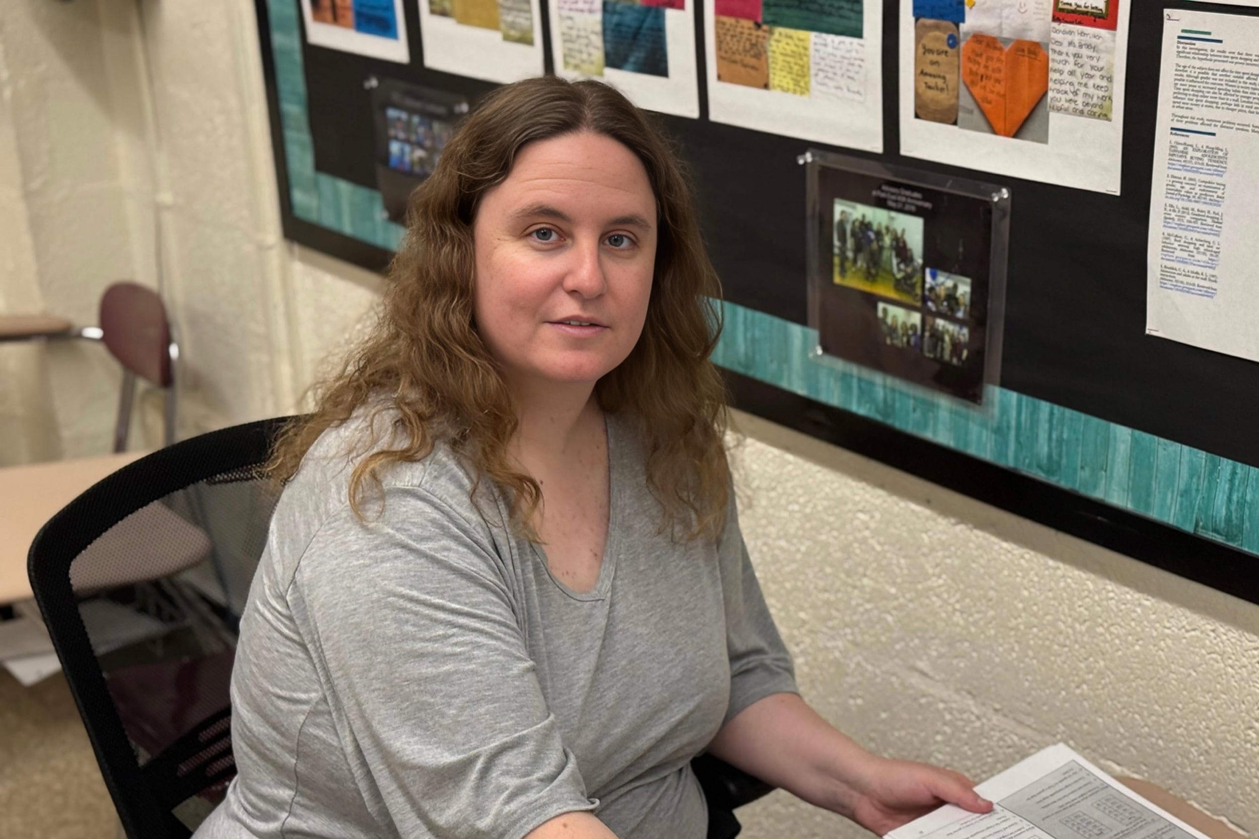 A white woman with long light brown hair and wearing a grey shirt poses for a portrait sitting at a desk in a classroom.