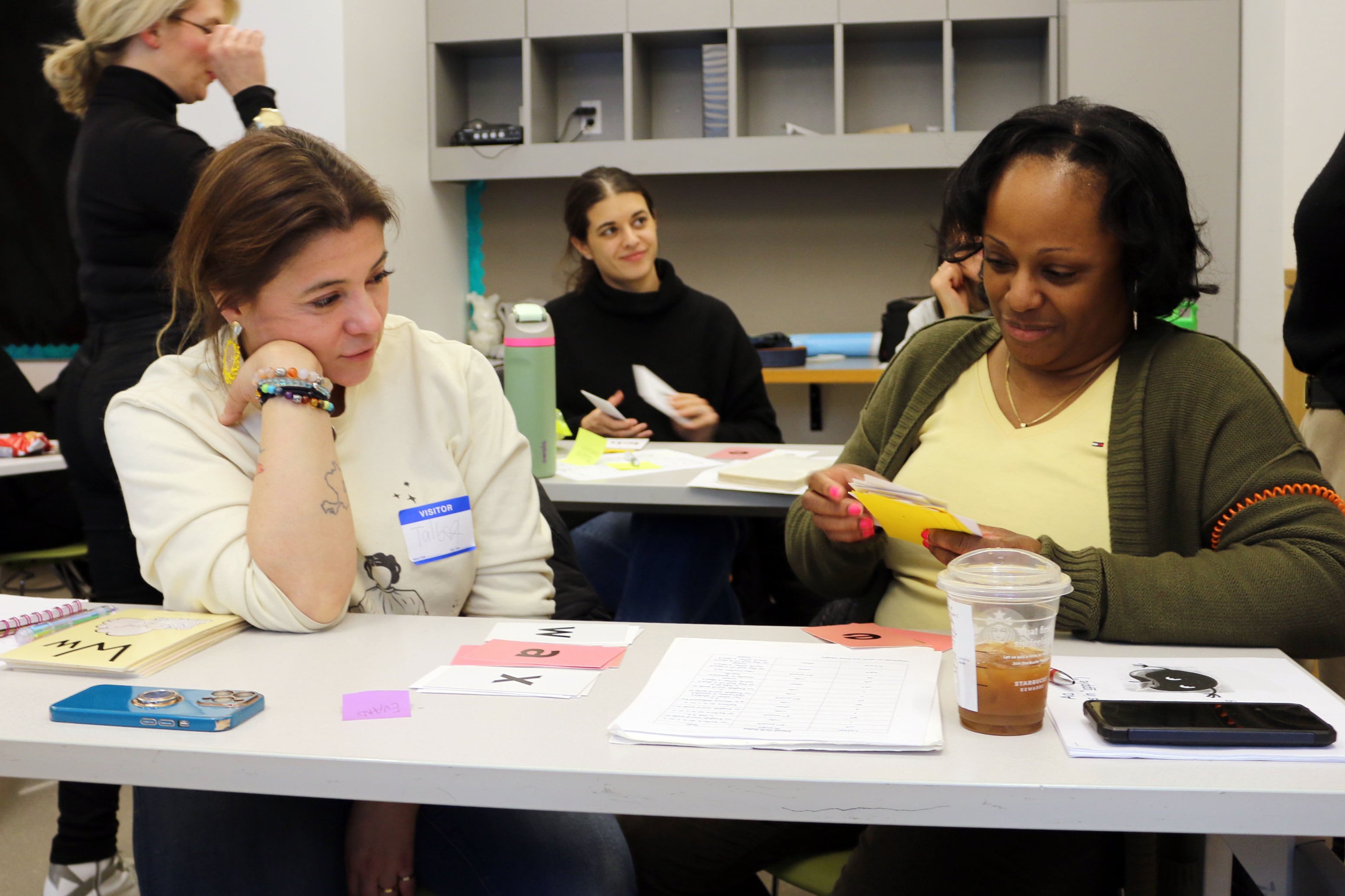 Two women sit at a table inside of a classroom practicing phonics training together with other people in the background.