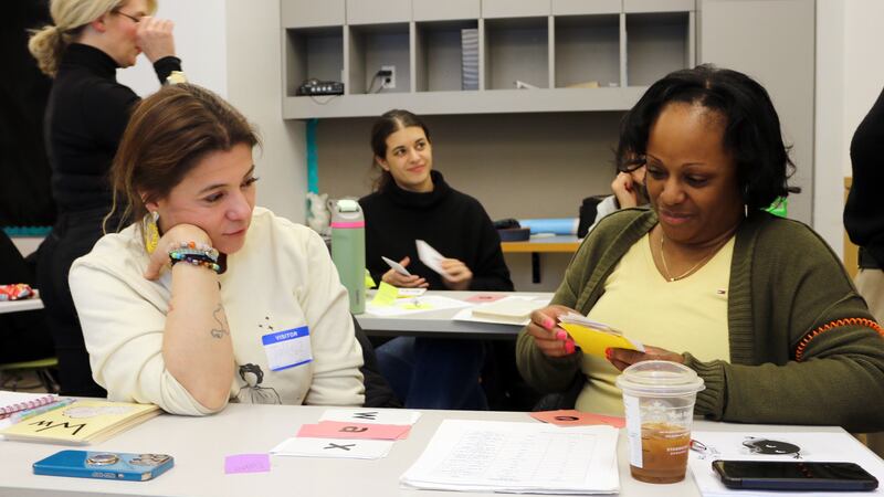 Two women sit at a table inside of a classroom practicing phonics training together with other people in the background.