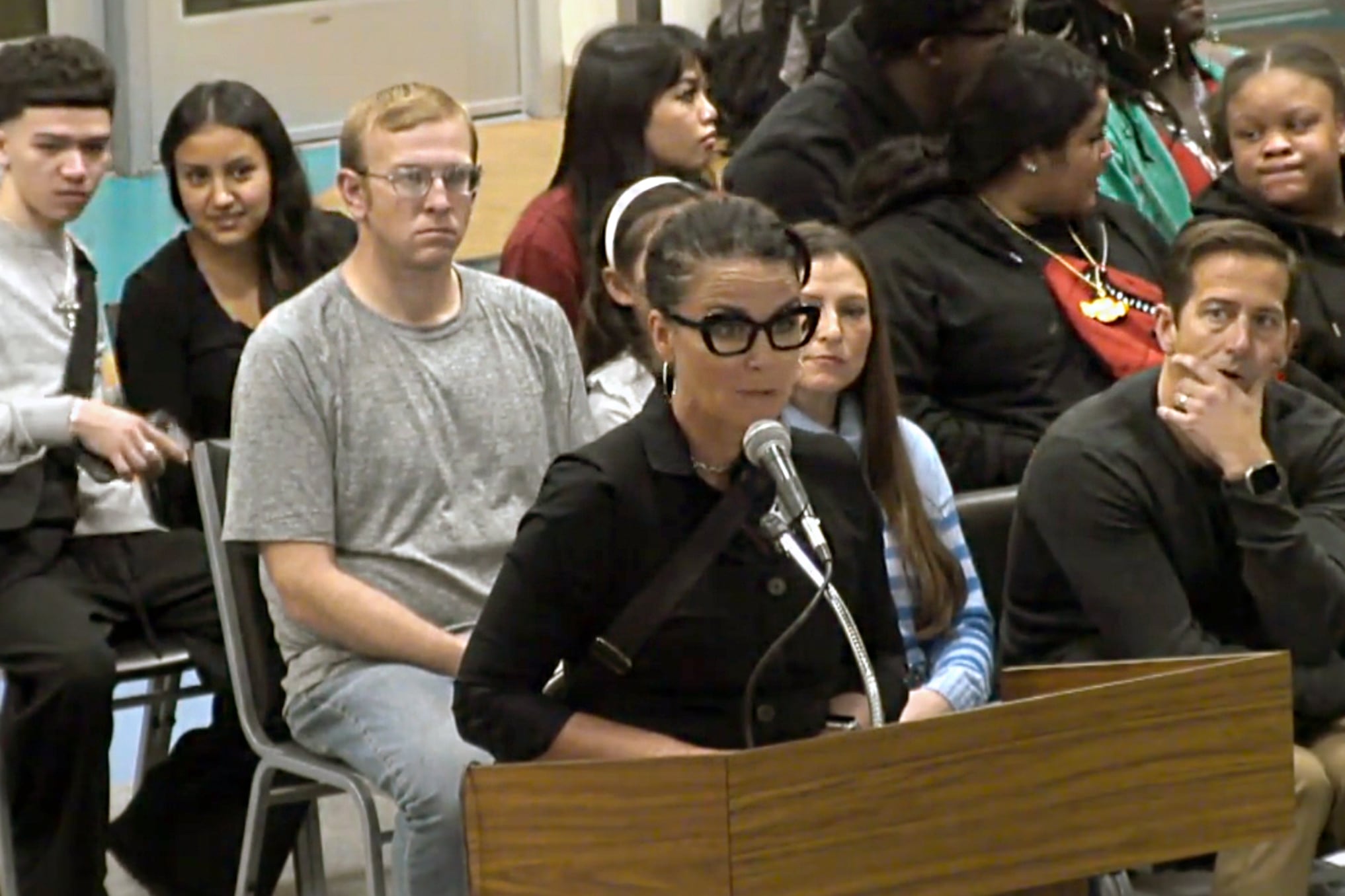 A woman with black glasses and a black shirt stands at a podium speaking into a microphone while a crowd of people sit in chairs in the background.