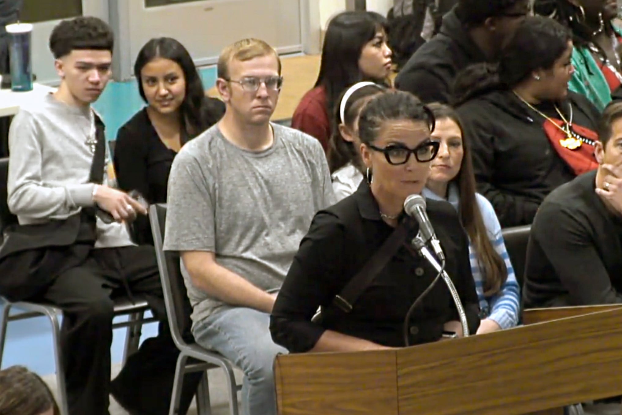 A woman with black glasses and a black shirt stands at a podium speaking into a microphone while a crowd of people sit in chairs in the background.