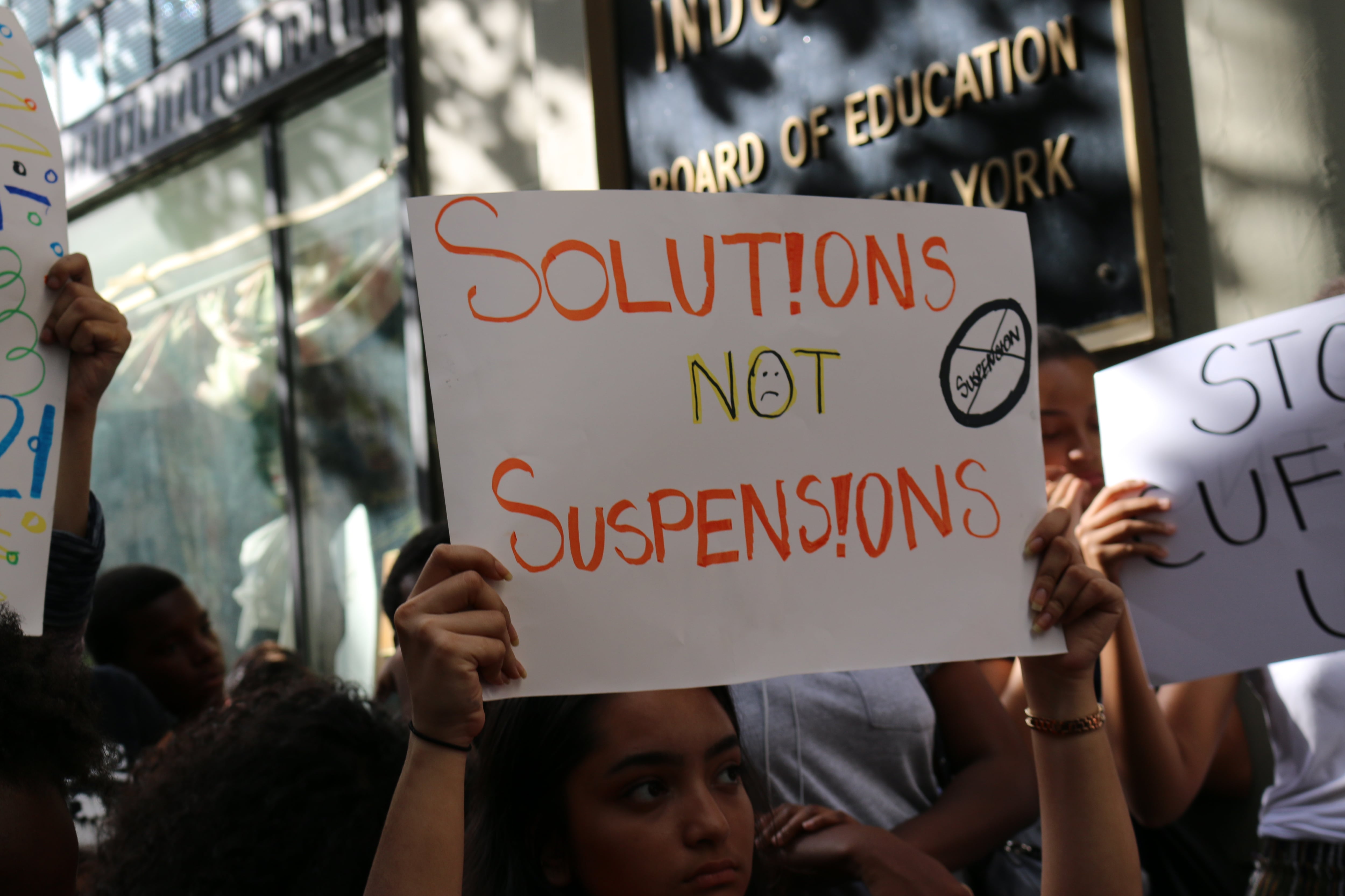 A person holds up a sign that reads "Solutions not suspension," in a crowd of people that you can't see. Board of Education building is in the background.