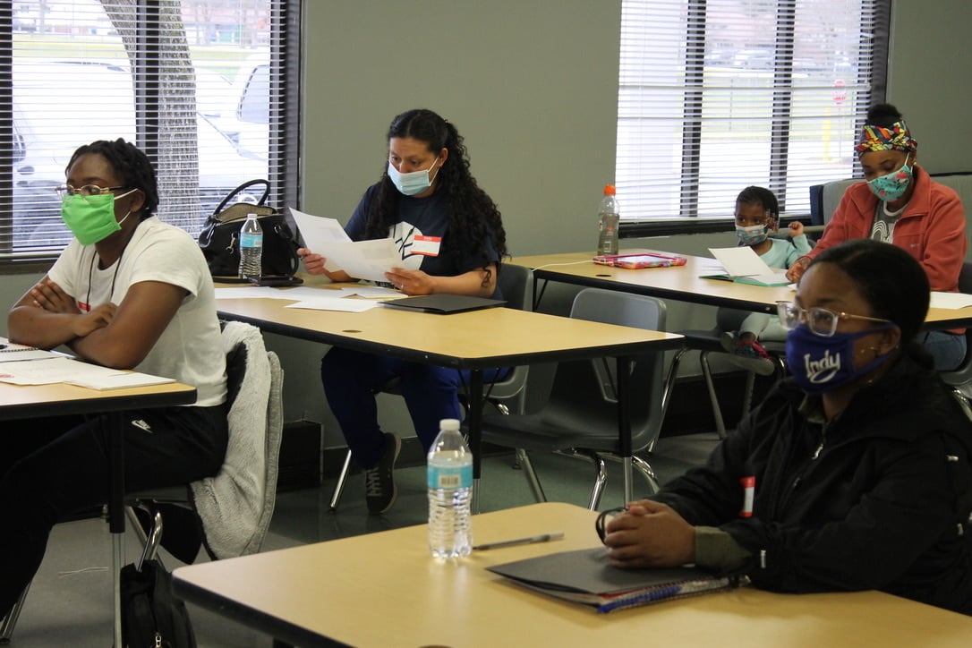 Four adult students wearing masks sit at spaced out desks in a classroom. One student has a small child next to her.