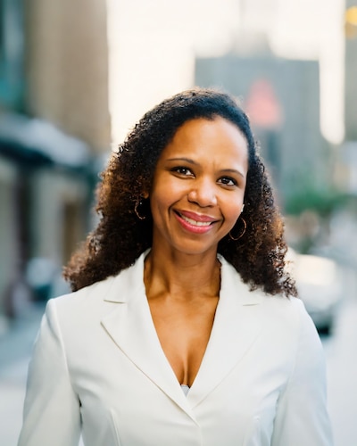 Headshot of a woman with dark, curly hair wearing a white blazer.