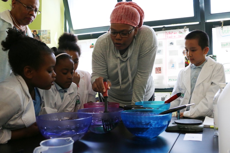 A Black adult teaches a group of young Black students all looking at an experiment on a classroom table.