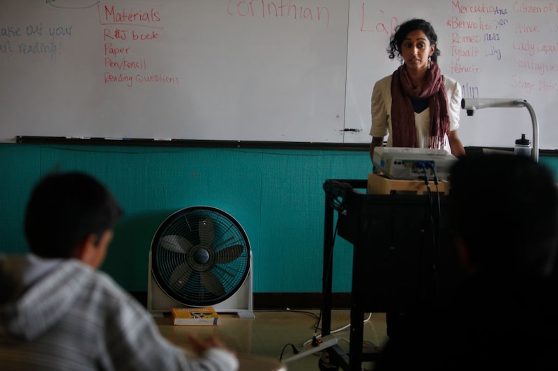 A student teacher stands at the front of a classroom behind a projector and students at desks and in front of a dry erase board.