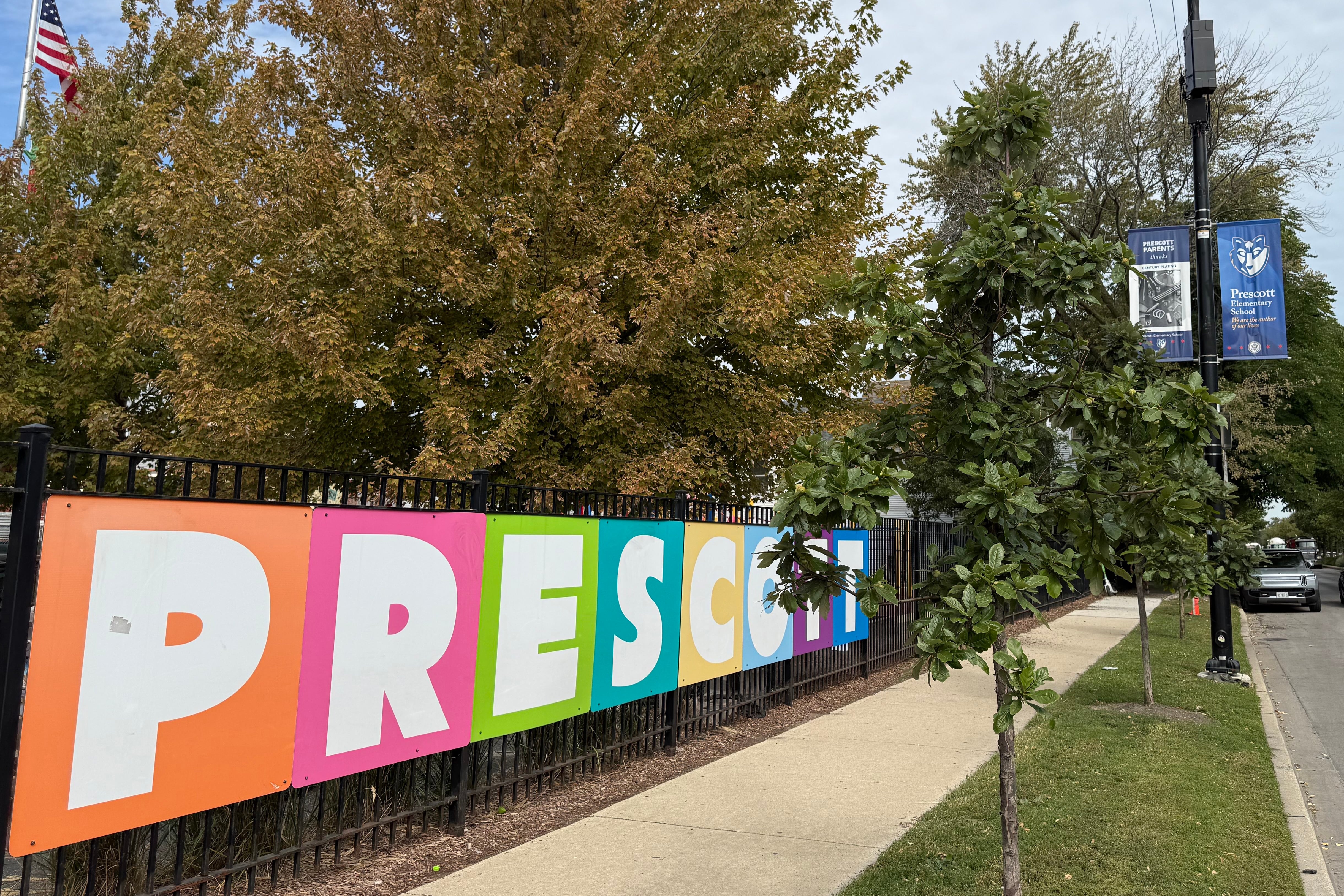 A photograph of a large colorful sign on a black metal fence that reads "Prescott" on a sunny day outside.