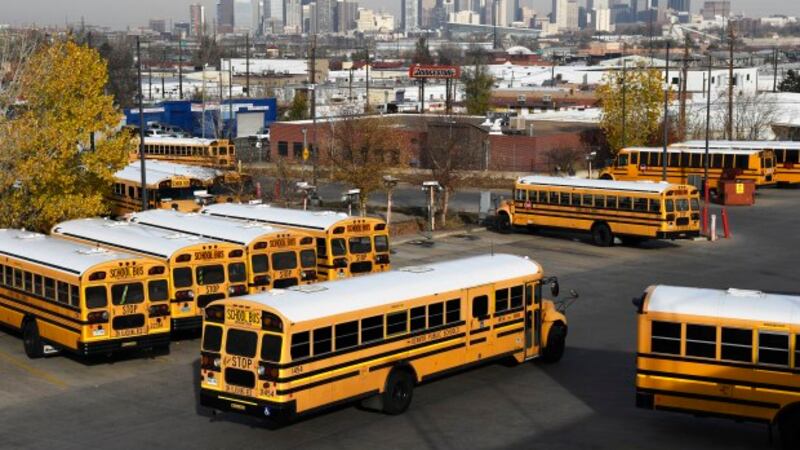 Yellow school buses head out past other parked buses in a terminal lot.