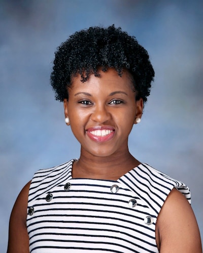 A photograph of a Black woman with short dark hair and wearing a black and white blouse and poses for a portrait against a blue background.