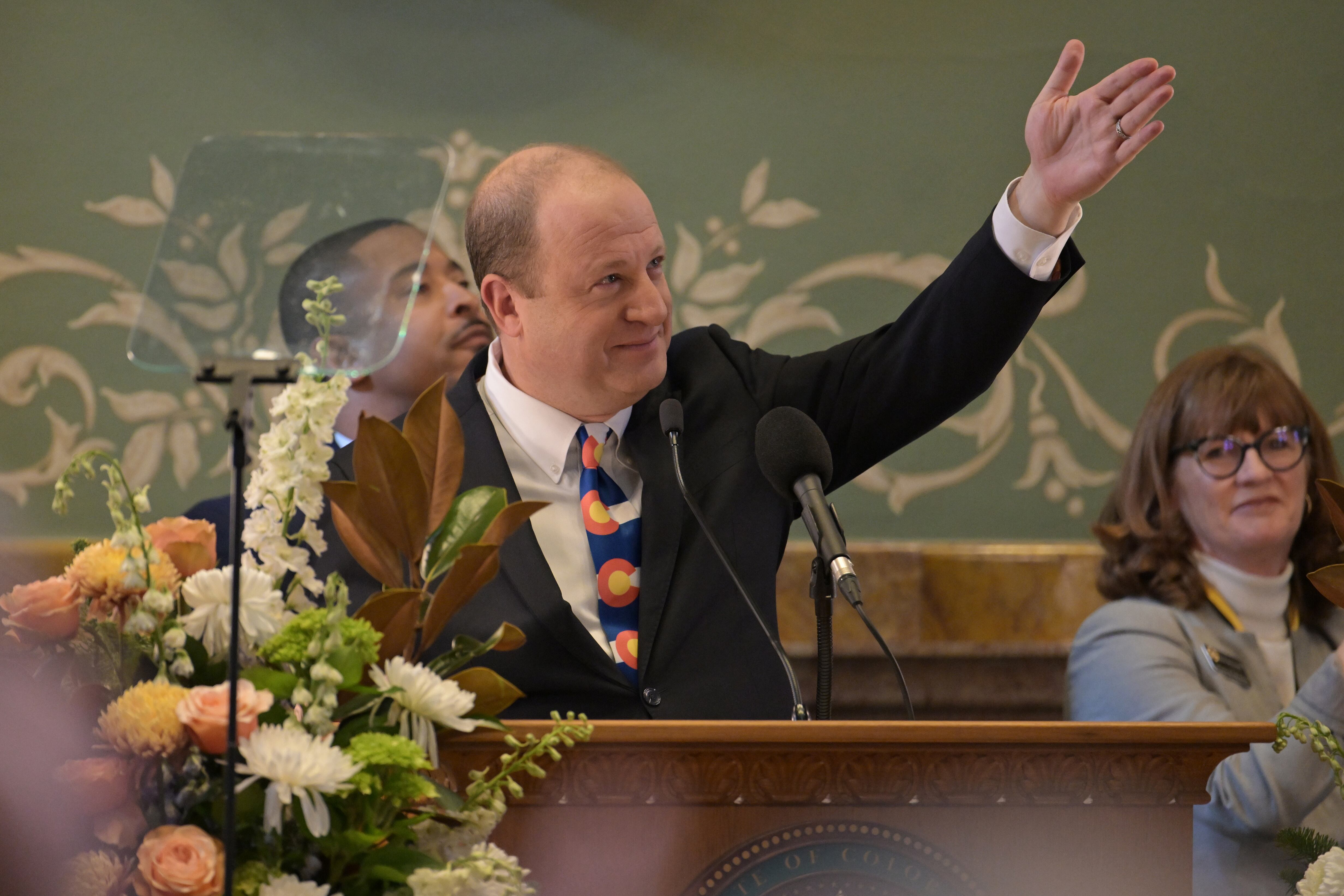 A man in a suit stands with one arm in the air and behind a wooden podium and in front of a green wall and two people sitting in the background.