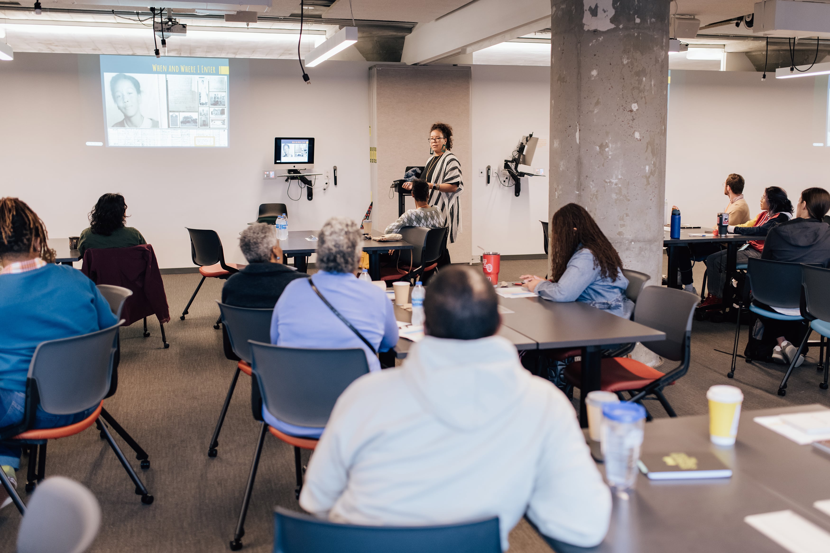 Several people are seated with their backs to the camera as they watch a woman in a black and white sweater present at the front of the room.