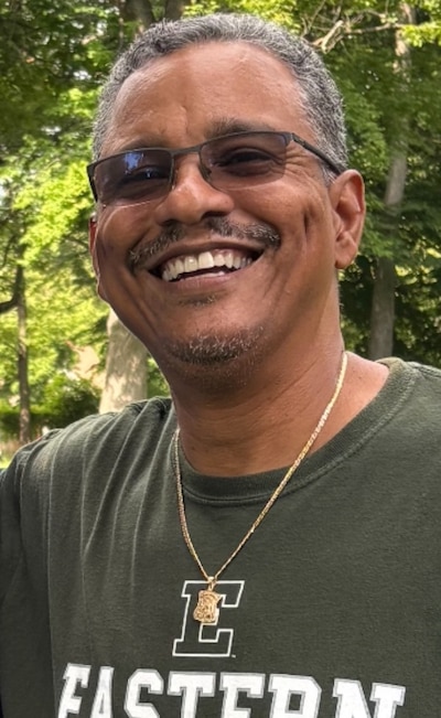 A man with short dark hair wearing glasses and a green shirt poses for a portrait outside.