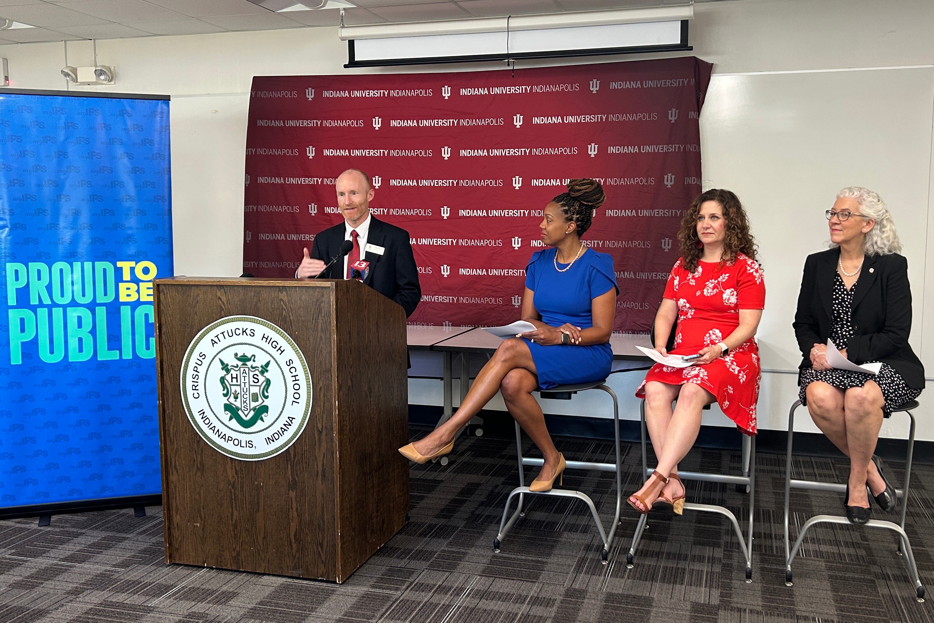 A man stands behind a podium with an emblem reading “Crispus Attucks High School” speaking into a microphone. To the right are three women sitting in chairs looking on.