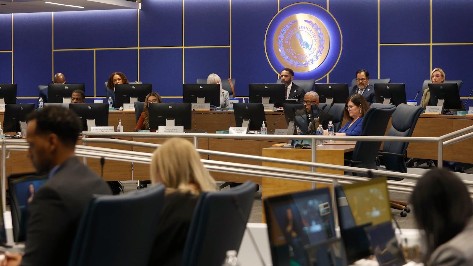People in business clothes sit in a large conference room with a blue wall and an illuminated circular sign in the background.