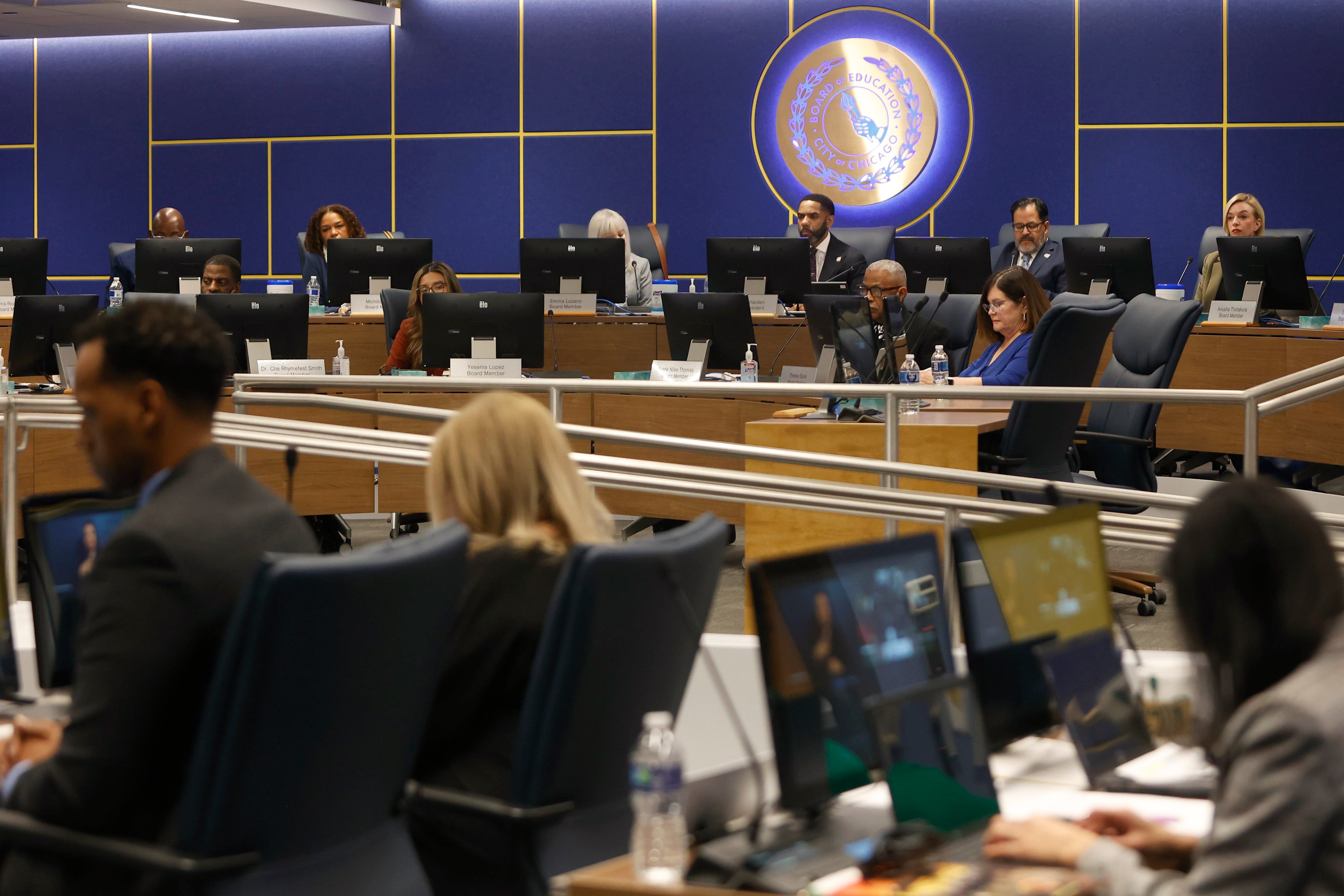 People in business clothes sit in a large conference room with a blue wall and an illuminated circular sign in the background.
