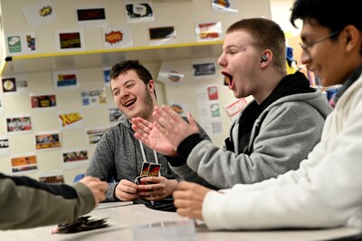 Three high school students sit at a table playing a card game with a decorated wall in the background.