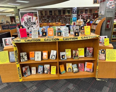 A bookcase in a school library has several banned books on display. It's draped in yellow caution tape.