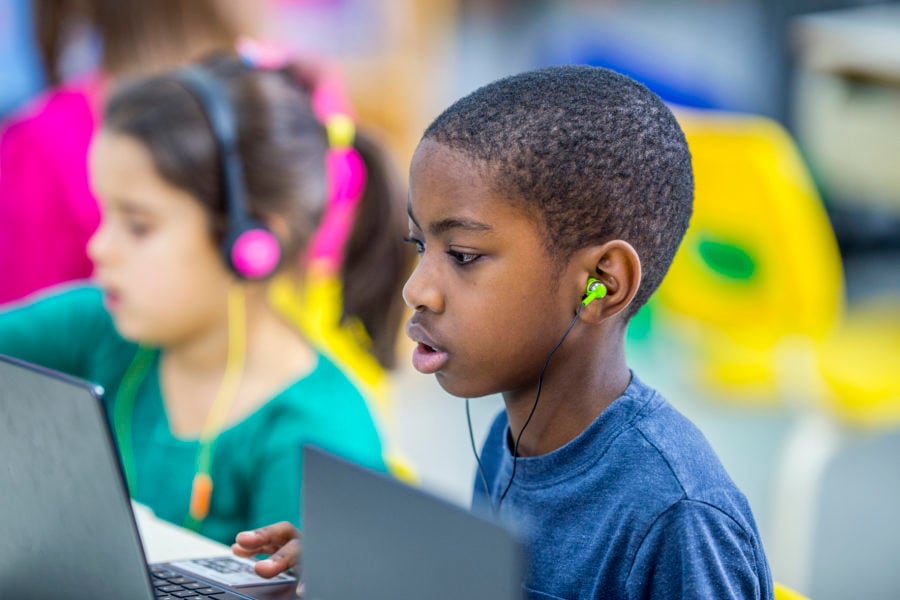 Two young students wearing headphones sit next to each other while they work on computers in a classroom.