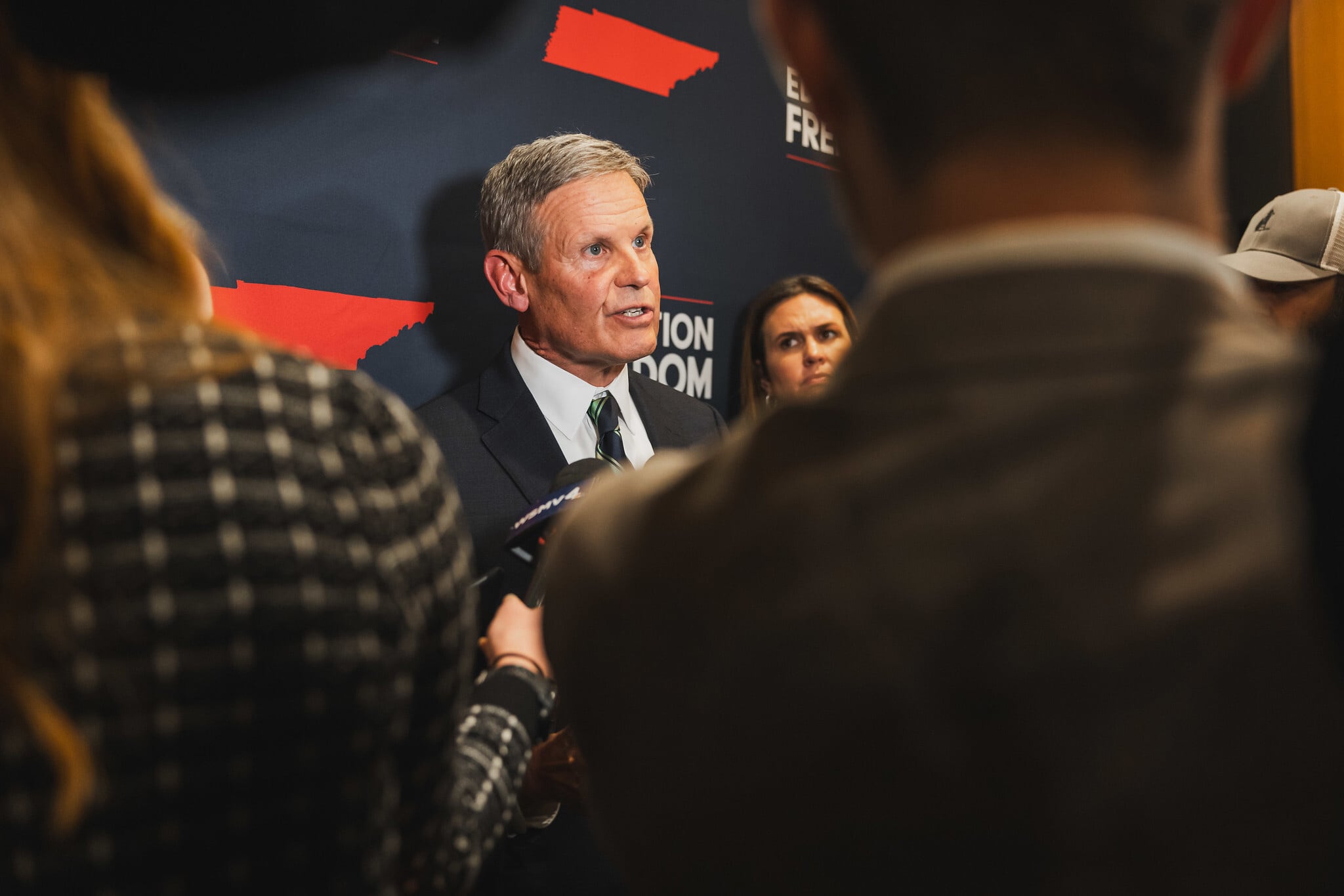 A man with short hair and wearing a dark suit is seen between people standing in front of him during a press conference.