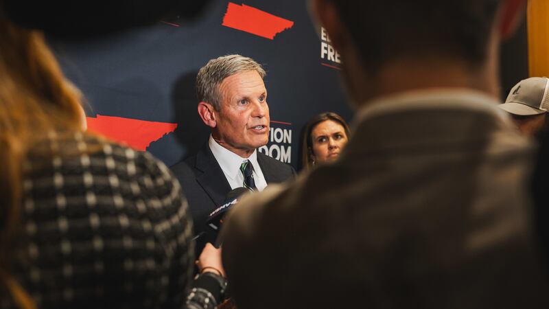 A man with short hair and wearing a dark suit is seen between people standing in front of him during a press conference.