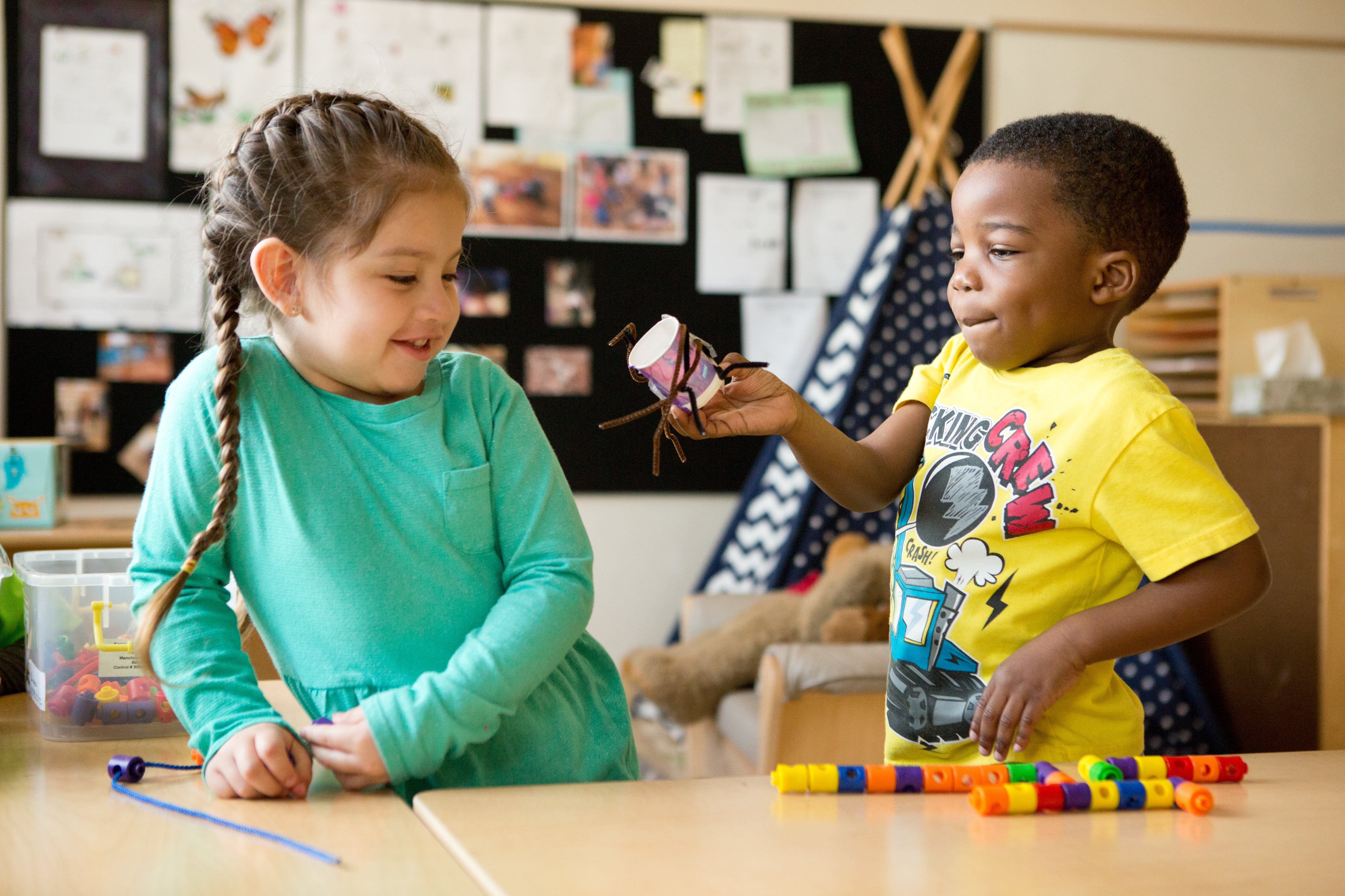 A preschool student shows his classmate a spider he made from pipe cleaners and a paper cup.