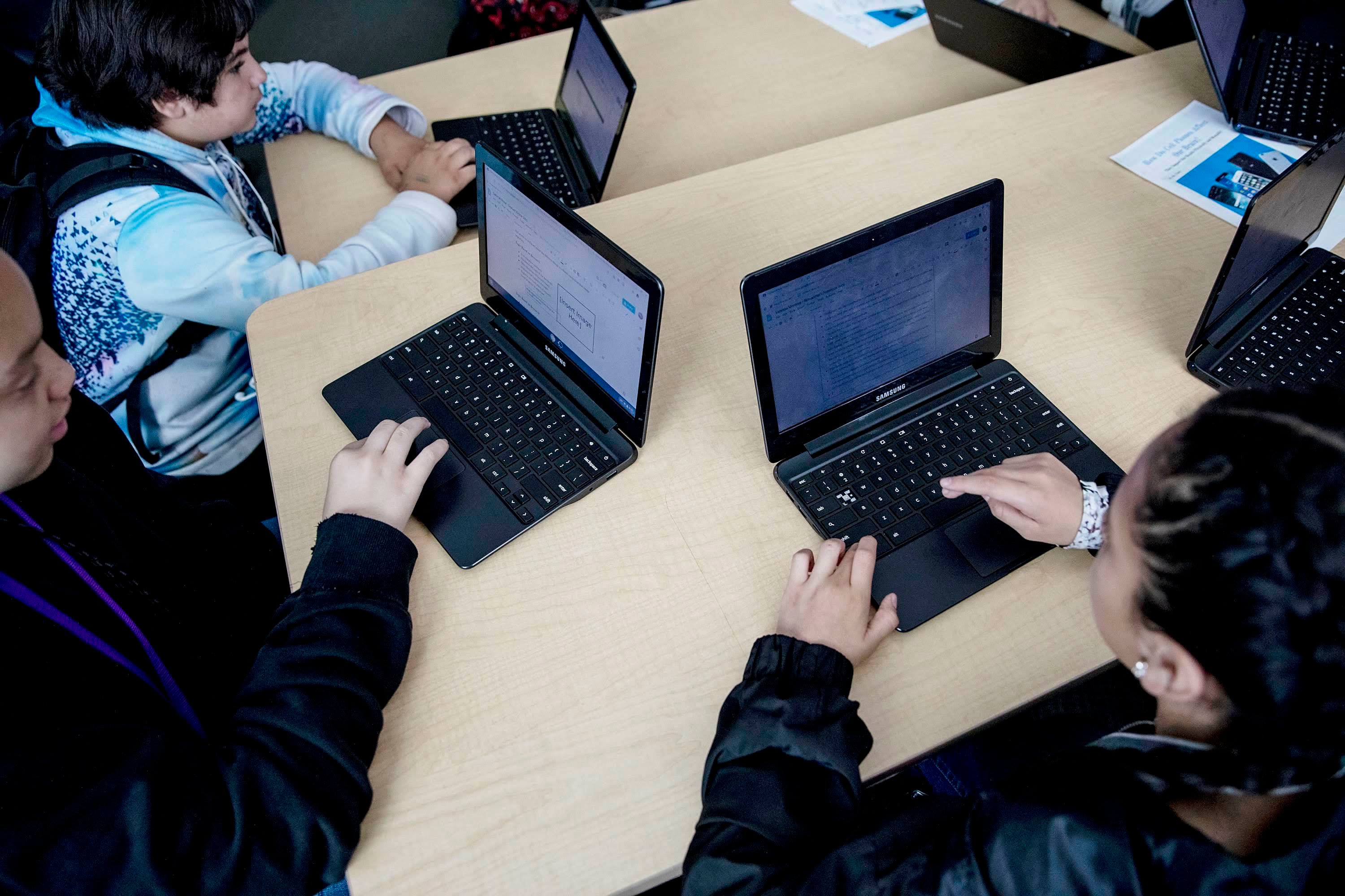 Three students work on laptops on a wooden desk. More laptops in the background.