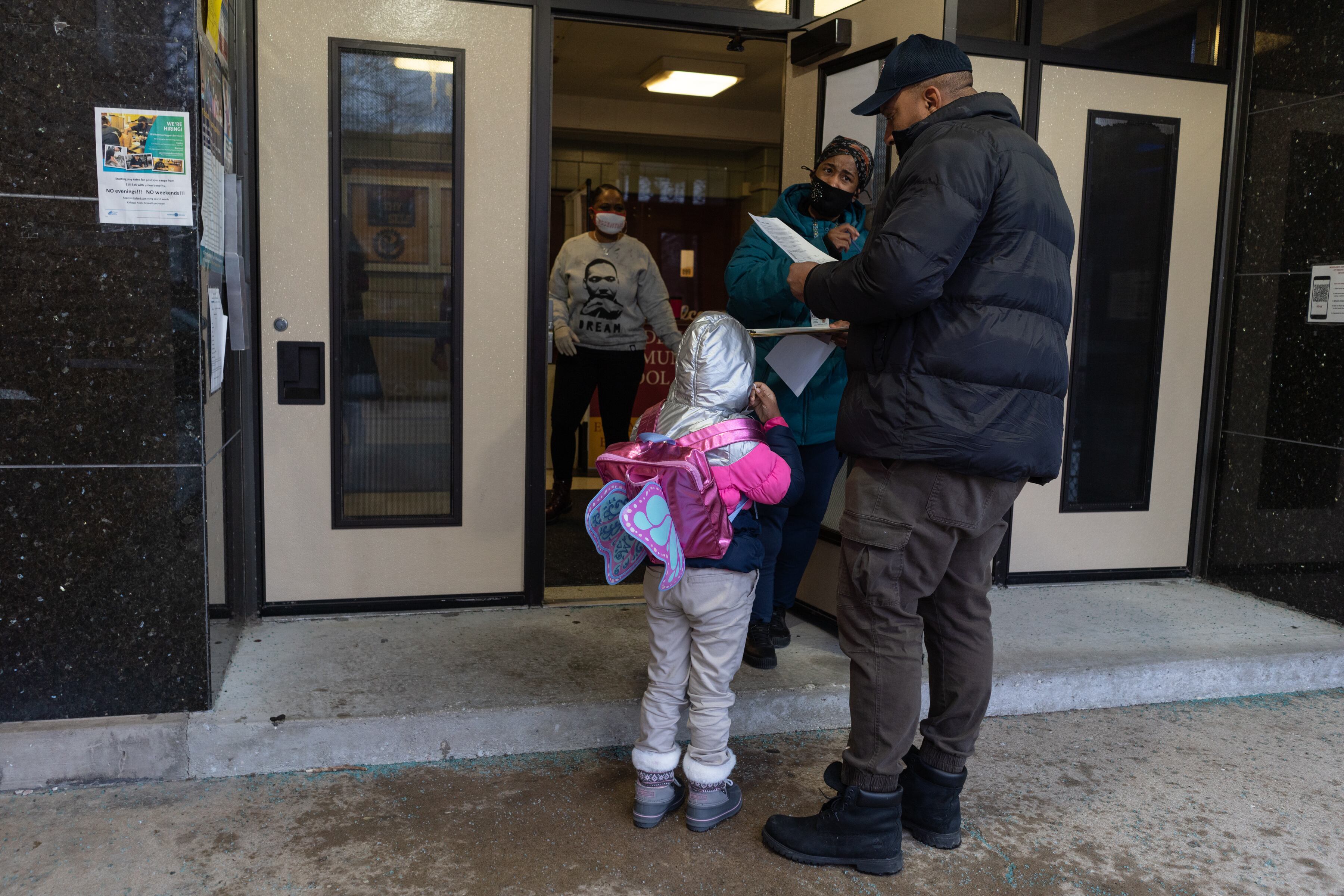 A father drops off his daughter at school. They are wearing winter clothing, and the girl is wearing a bright pink jacket and a butterfly backpack.