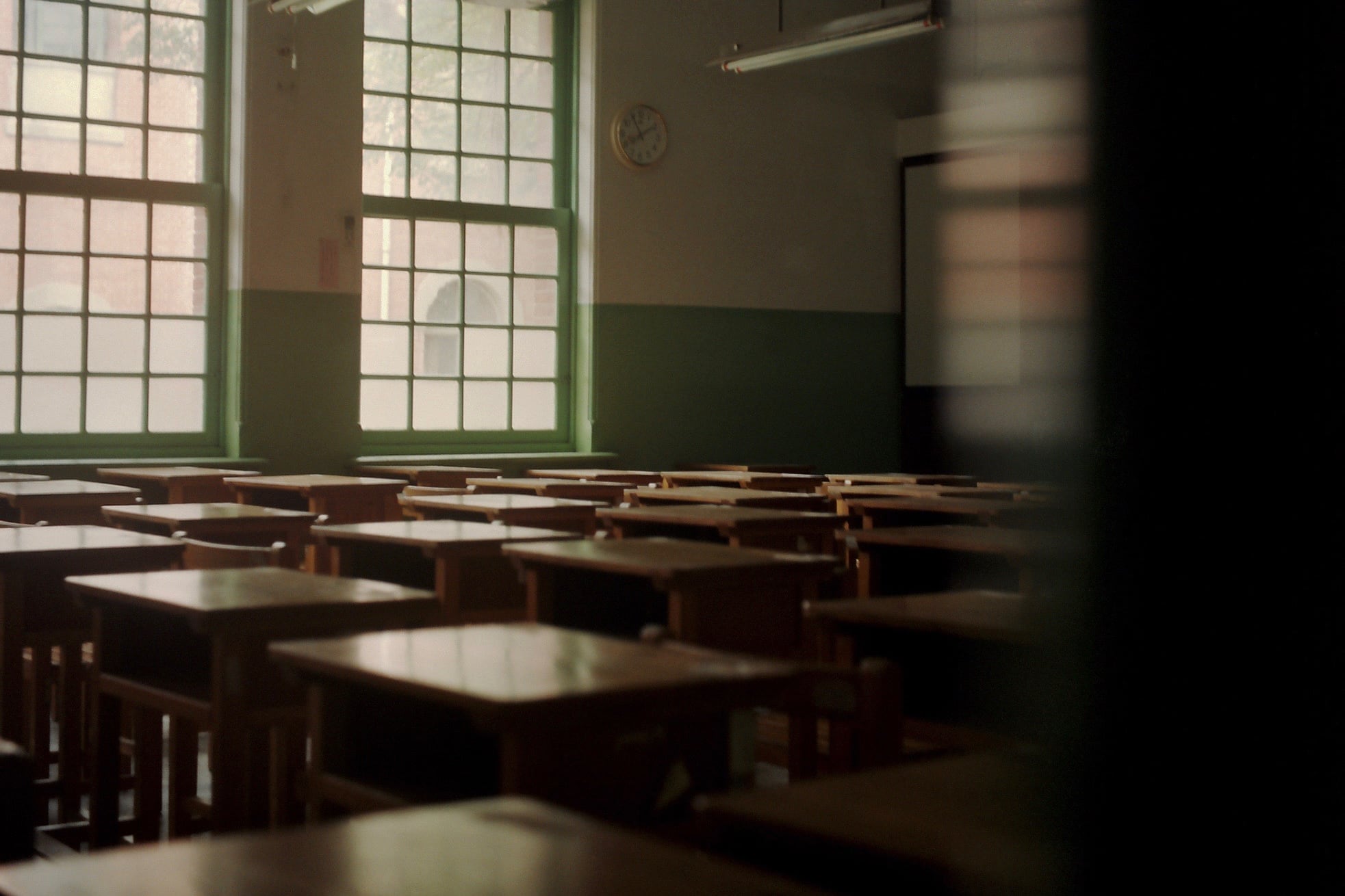 A dark empty classroom full of desks in rows with two windows on the left back wall.
