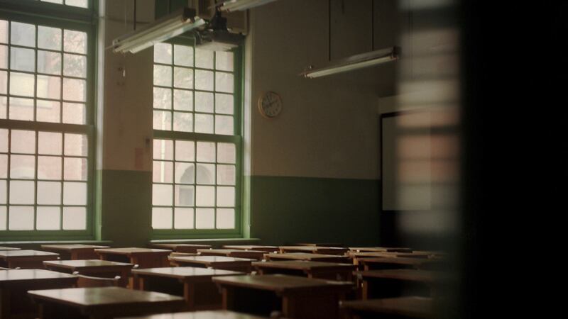 A dark empty classroom full of desks in rows with two windows on the left back wall.