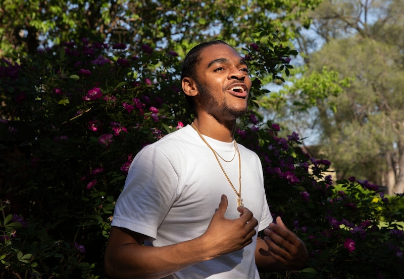A high school senior with dark hair and wearing a white t-shirt with a gold necklace with a diamond letter "K" poses for a portrait in front of a rose bush.