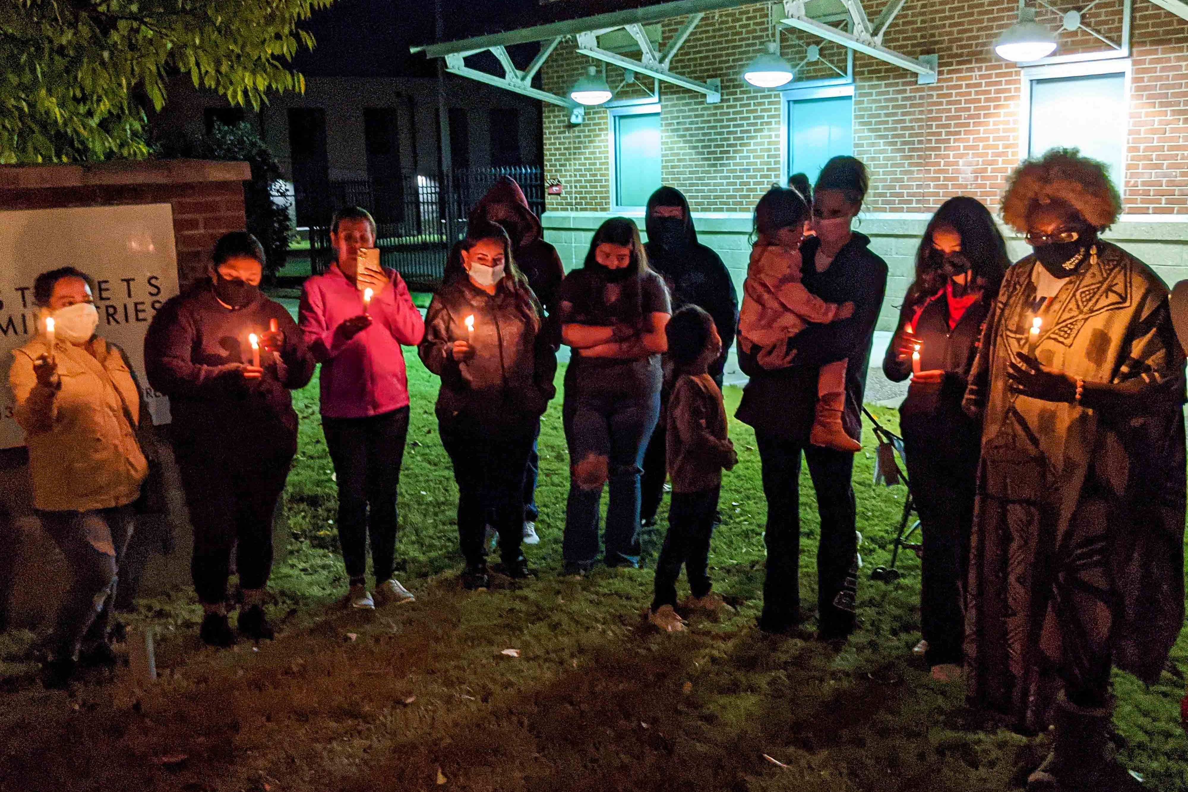 About a dozen people stand outside at night in a semi-circle holding candles during a candlelight vigil in front of a community center.