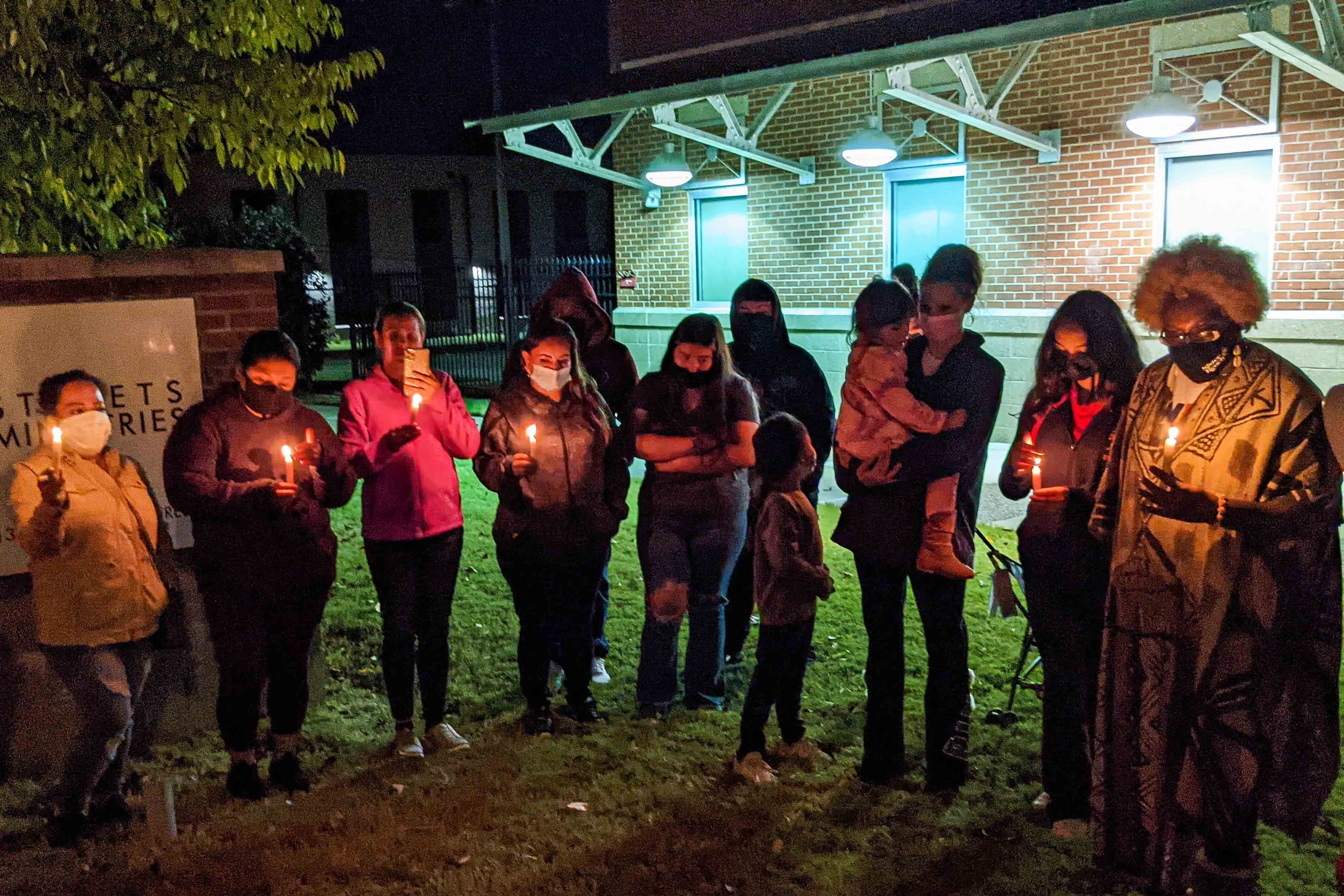 About a dozen people stand outside at night in a semi-circle holding candles during a candlelight vigil in front of a community center.