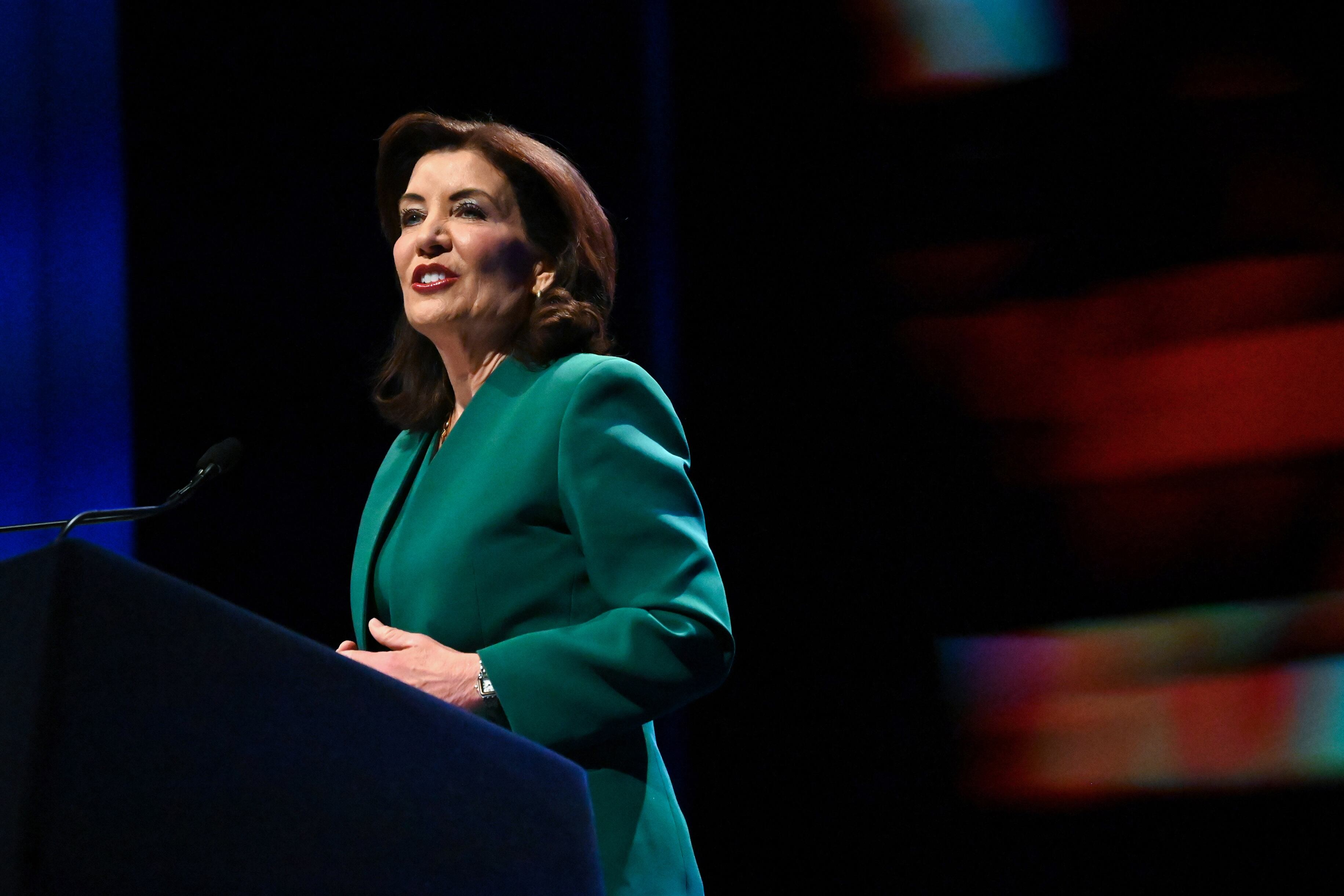 A woman with short dark hair wearing a green suit jacket speaks from behind a podium.