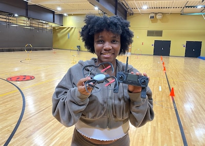 A girl in a gray sweatshirt holds a drone and another piece of equipment in a gymnasium.
