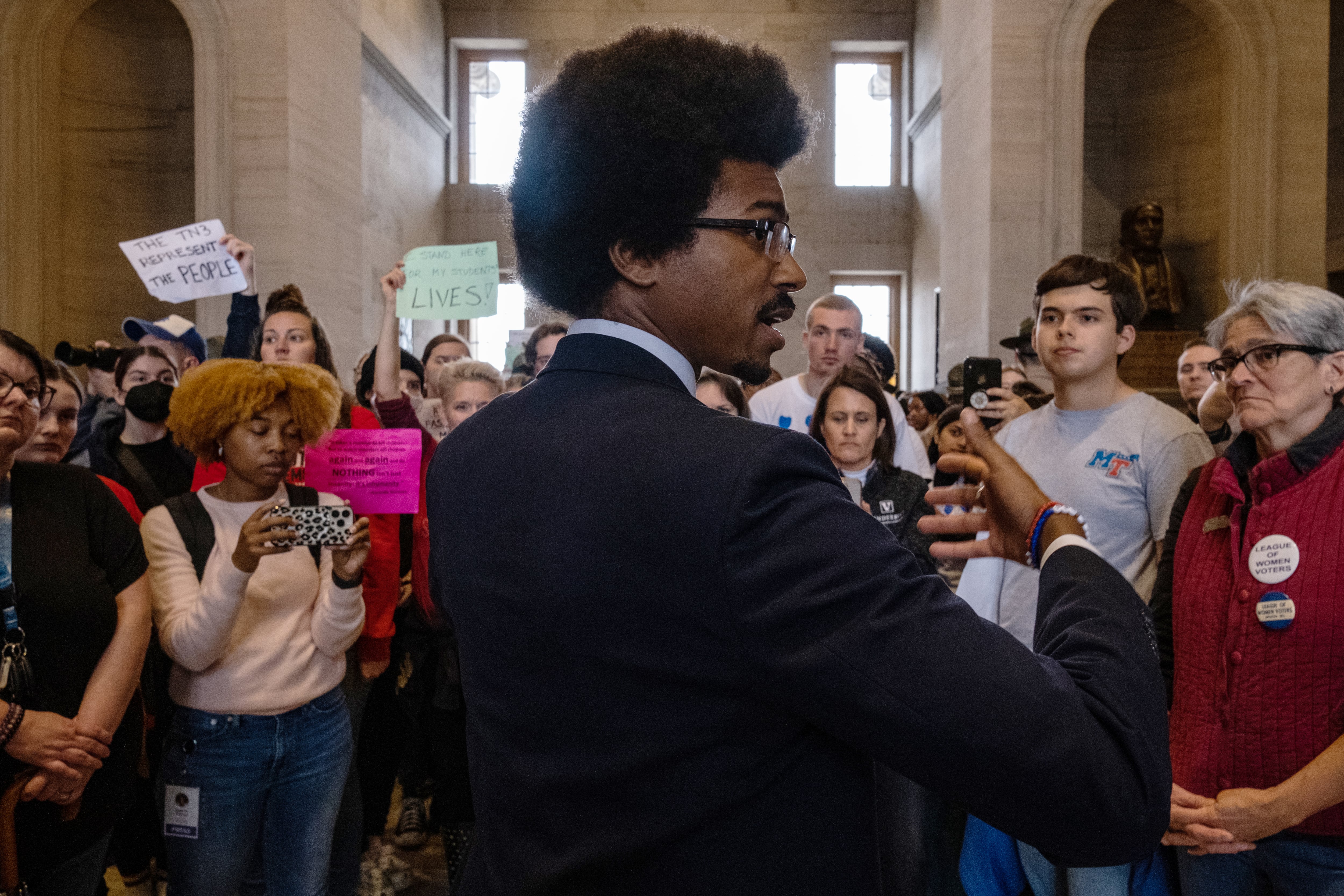 A man is turned mid-conversation with his right-side profile to the camera as many demonstrators look on in the distance.