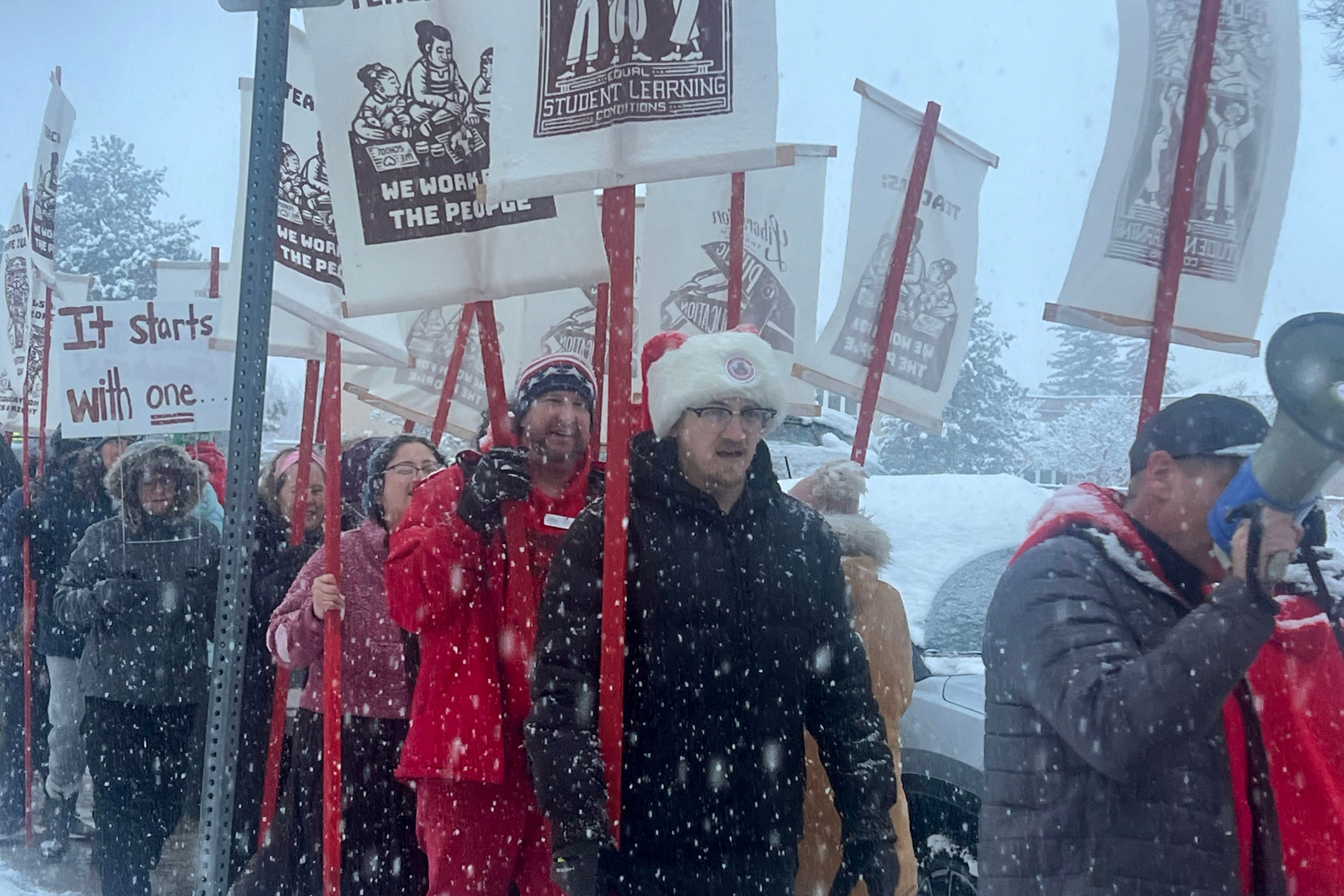 A photograph of protestors wearing heavy jackets and cold weather gear holding signs and marching in front of large building. It's snowing out and the sun is setting.