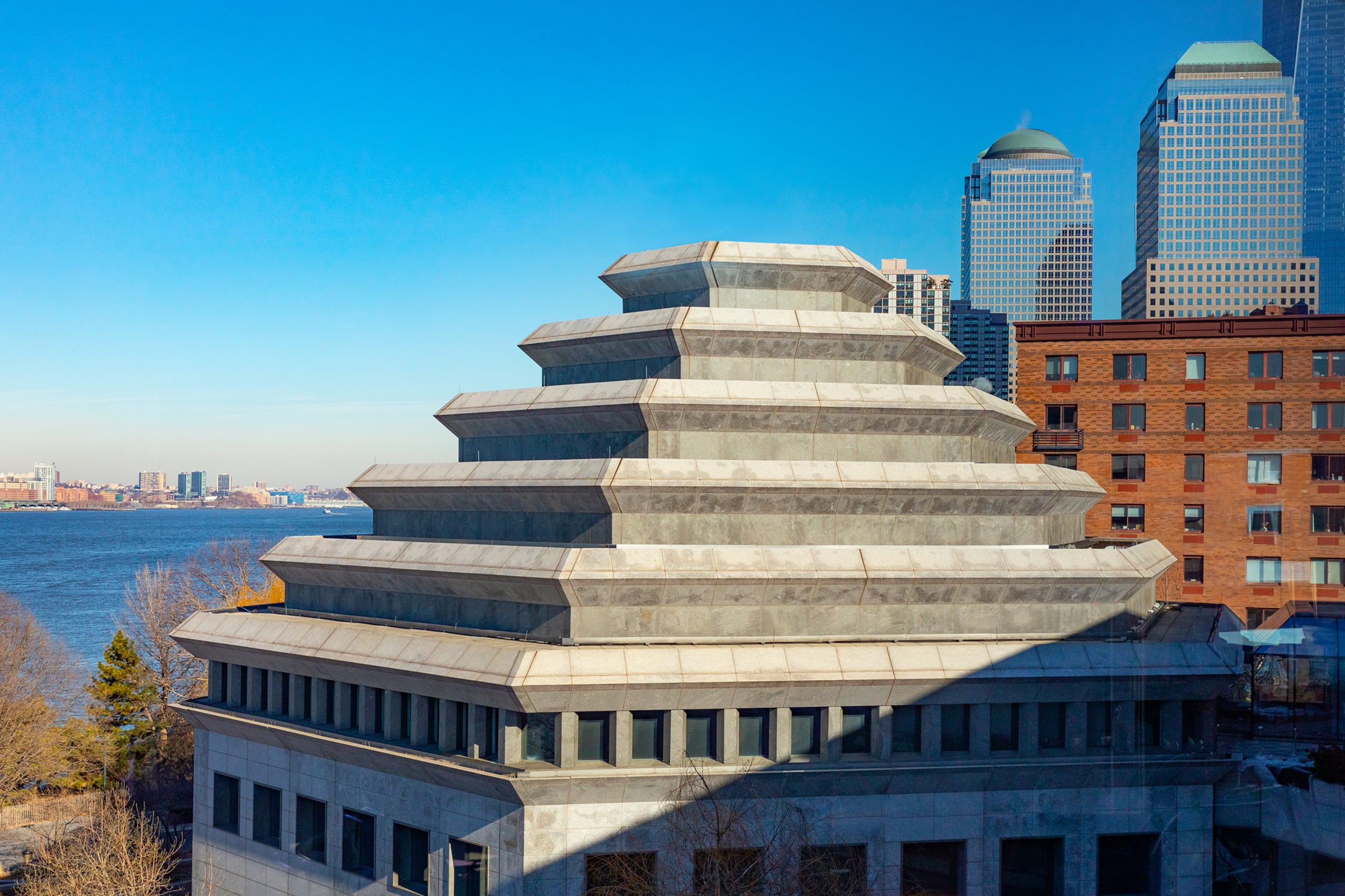 The top of a square building with a pyramid like shape against a blue sky with a river and other buildings in the background.