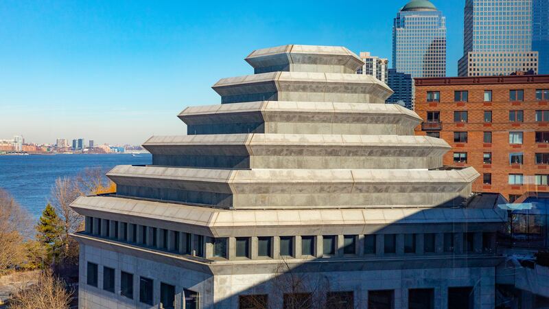 The top of a square building with a pyramid like shape against a blue sky with a river and other buildings in the background.