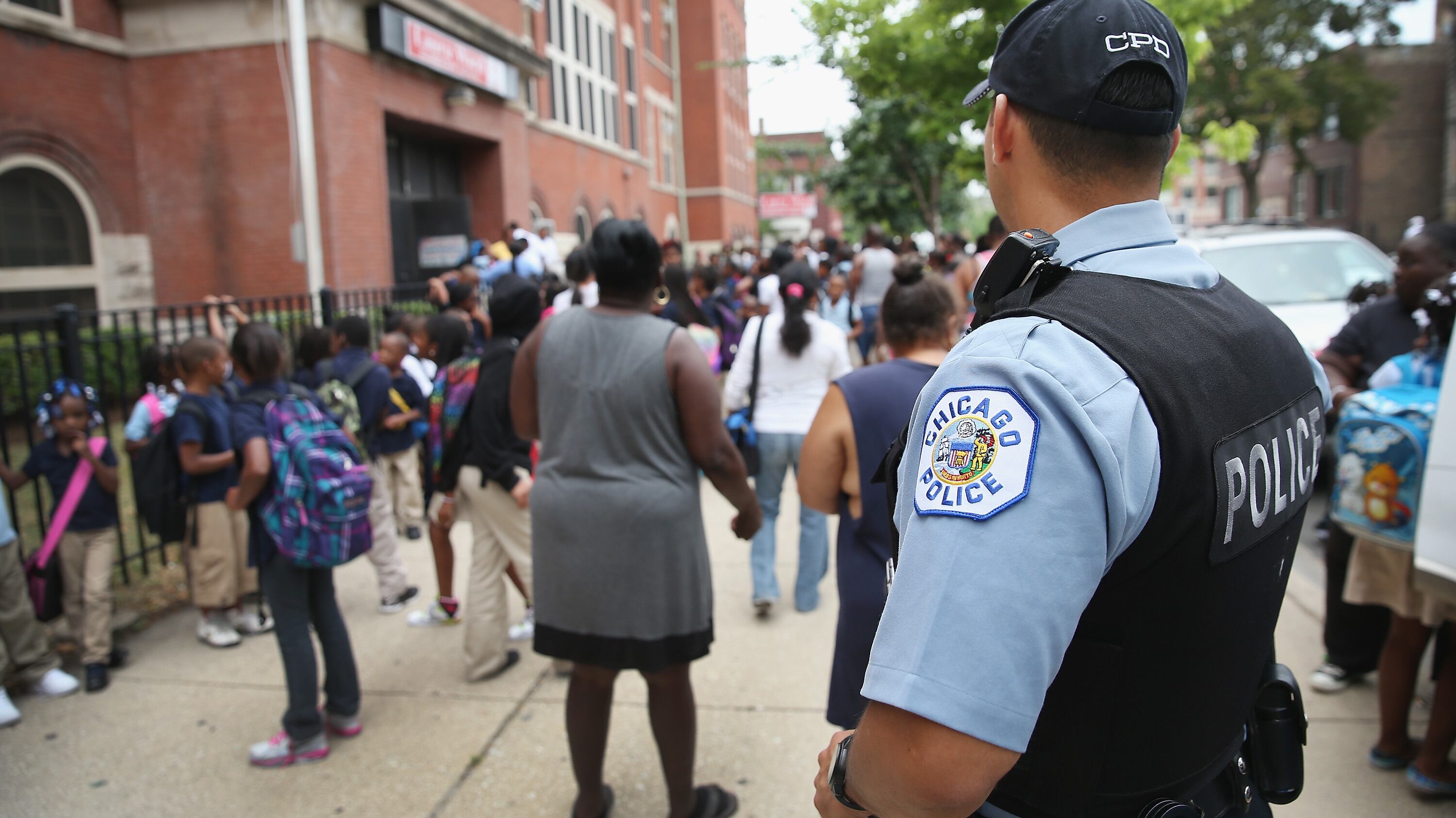 A police officer stands outside a school as students walk along the sidewalk.