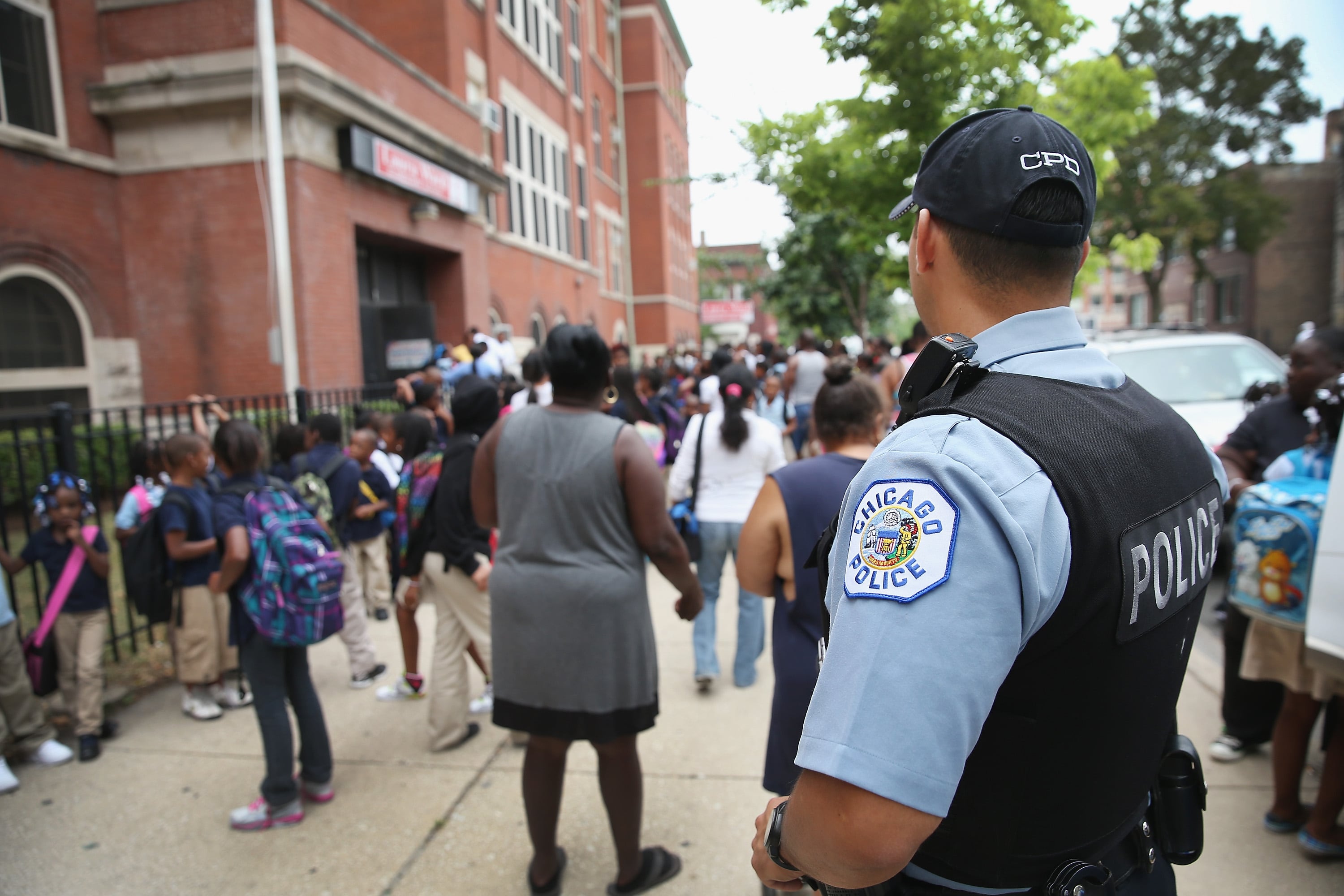 A police officer stands outside a school as students walk along the sidewalk.