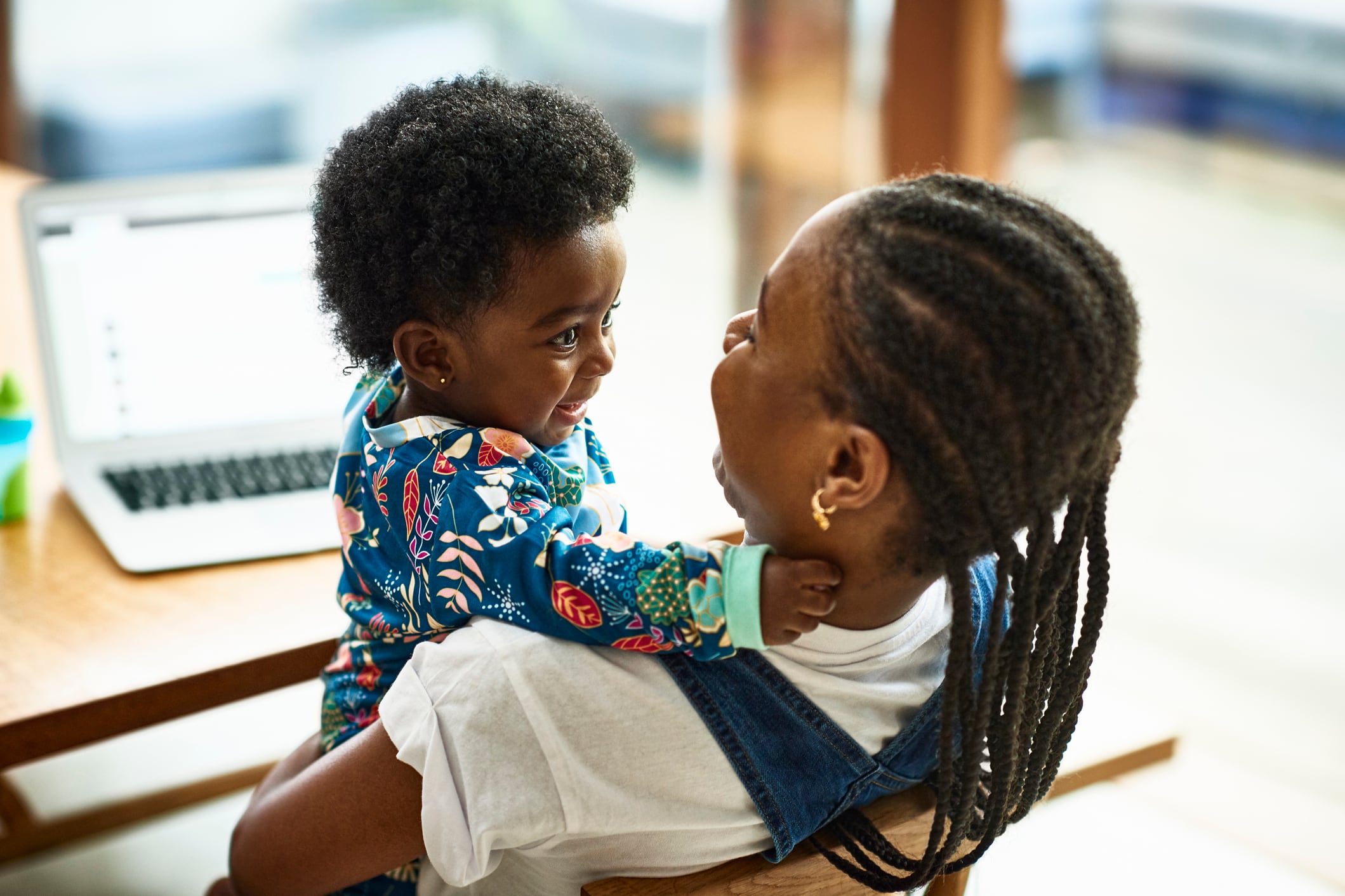 A photograph of a Black mother smiling and holding her small baby while sitting at a desk with her laptop in the background.