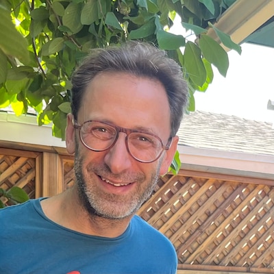 Headshot of a man with dark hair taken outside. He is wearing a blue shirt and glasses